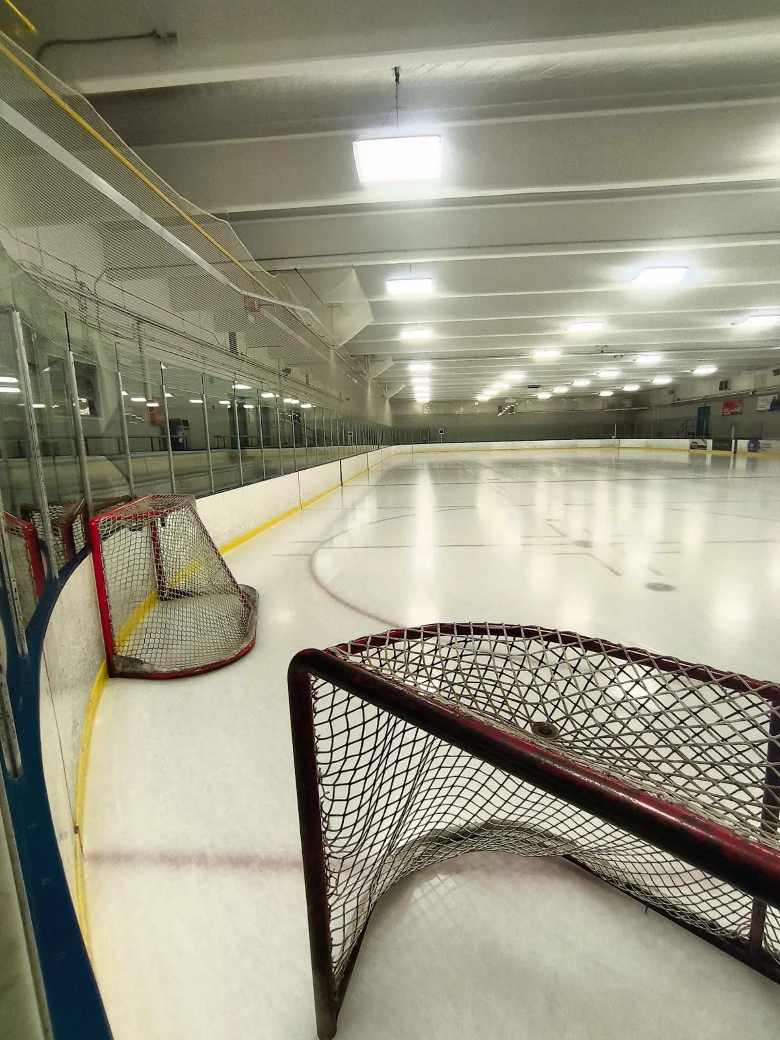 Empty indoor ice rink seen from ice level with two hockey nets in the foreground; full length of the ice sheet, boards, glass and overhead lighting visible, no people present. — Grand Trunk Arena