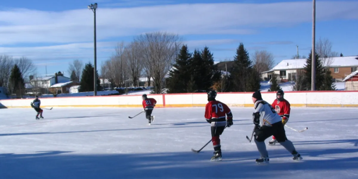 Players skating on the big outdoor rink at the Laurier-Station recreation complex, with floodlight poles, bare trees and houses beyond the boards. — Grande patinoire du Complexe Récréatif (Laurier-Station)