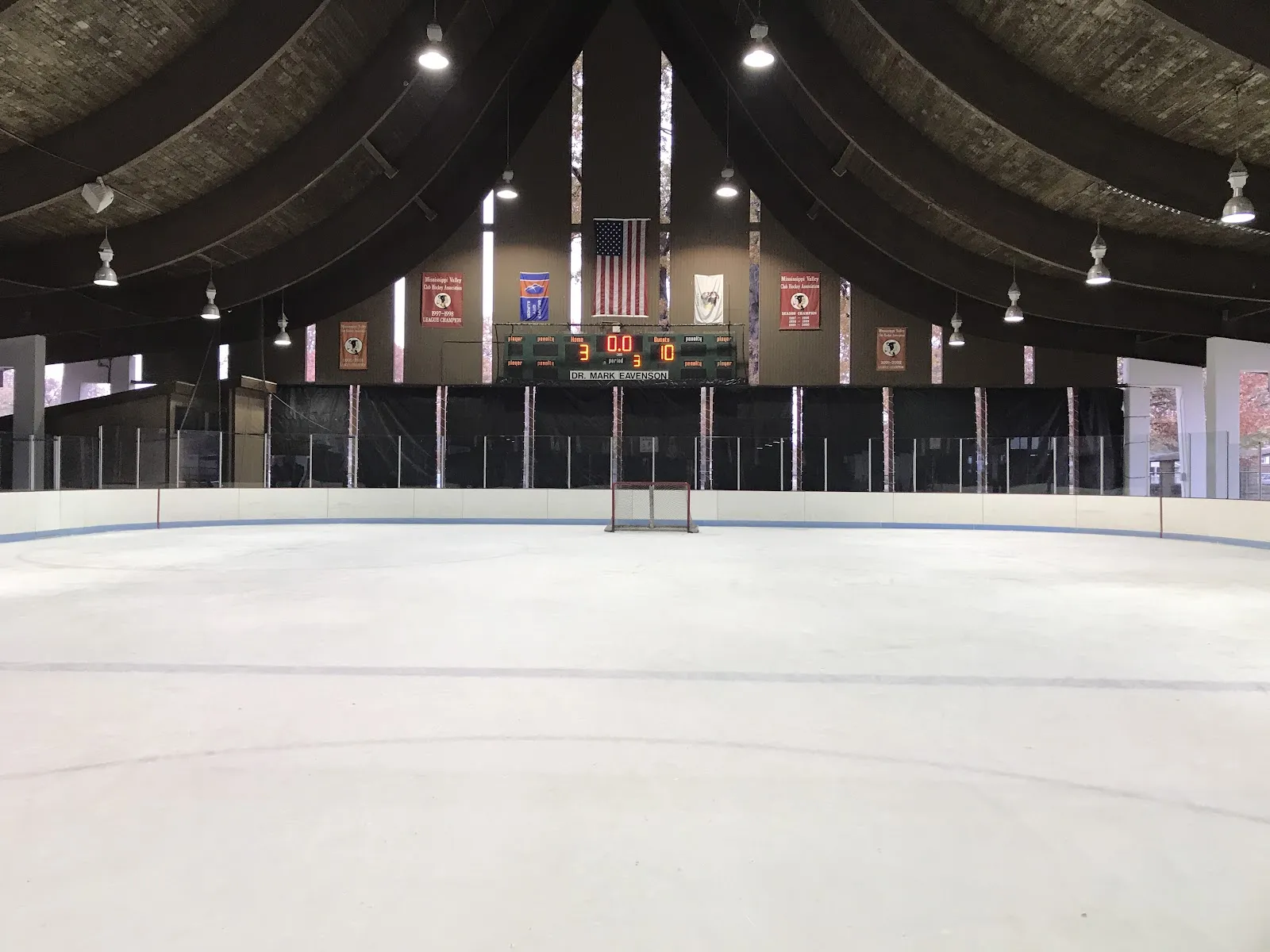 La pista de hielo vacía bajo el techo arqueado, vista desde un extremo, con bardas y vidrio a los lados y una portería, el marcador y una bandera en el extremo del fondo. — Granite City Ice Rink