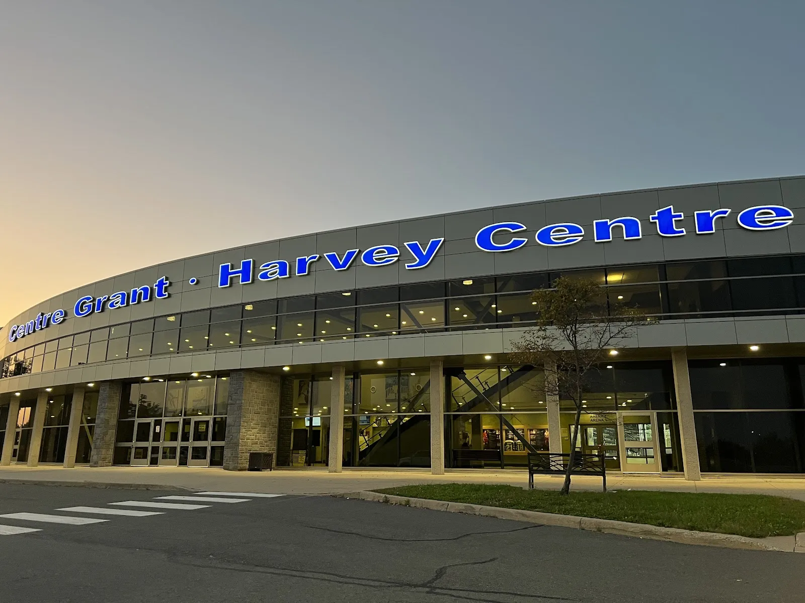 El Grant-Harvey Centre al atardecer, con su fachada curva de vidrio iluminada bajo las letras azules de Centre Grant-Harvey Centre — Grant-Harvey Centre