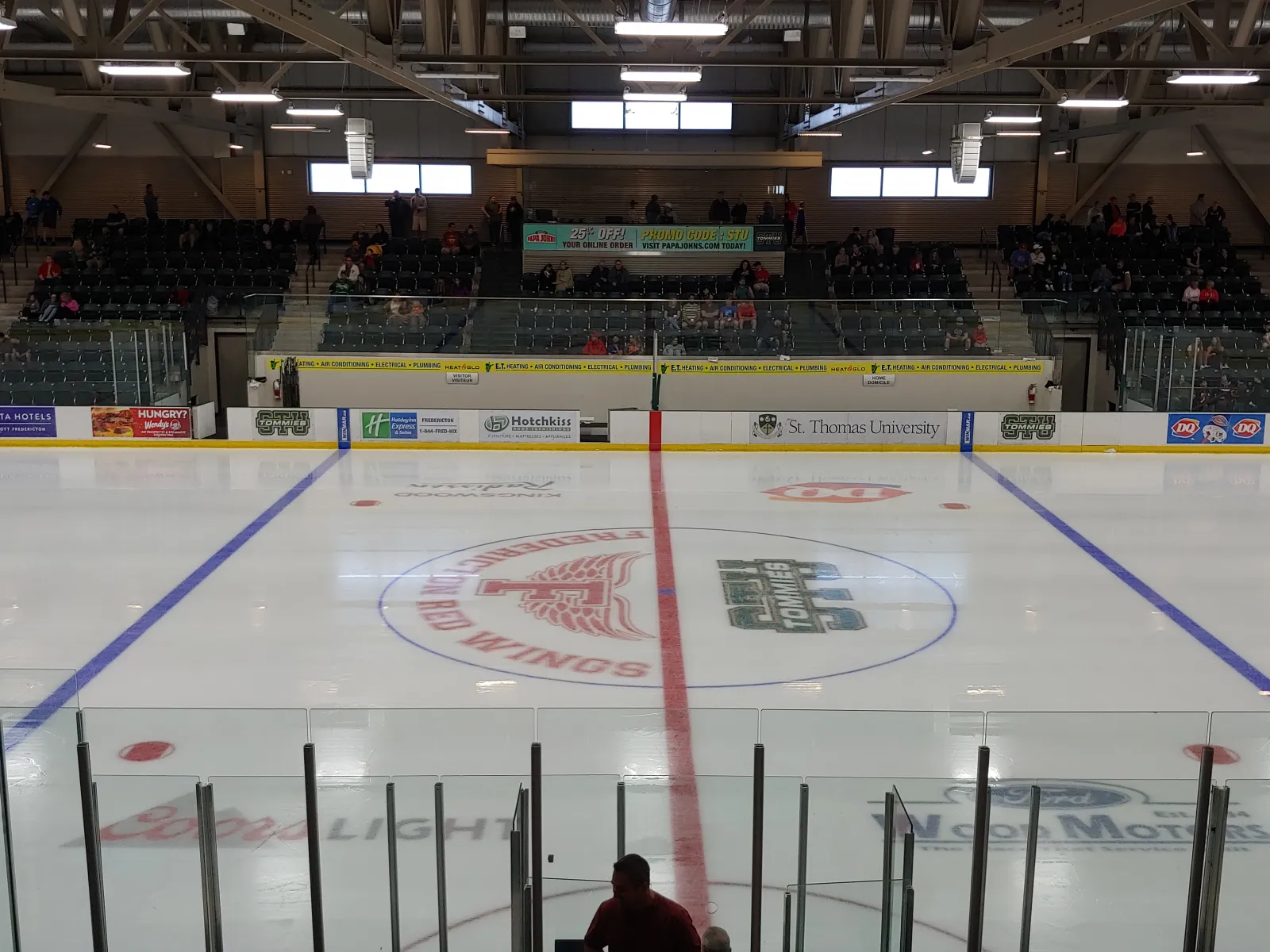 Vista a lo largo del hielo desde detrás de las vallas, con toda la pista, el cristal, las gradas con espectadores y el logotipo del centro de la pista que dice "FREDERICTON RED WINGS" con el logotipo de los STU Tommies a un lado — Grant-Harvey Centre