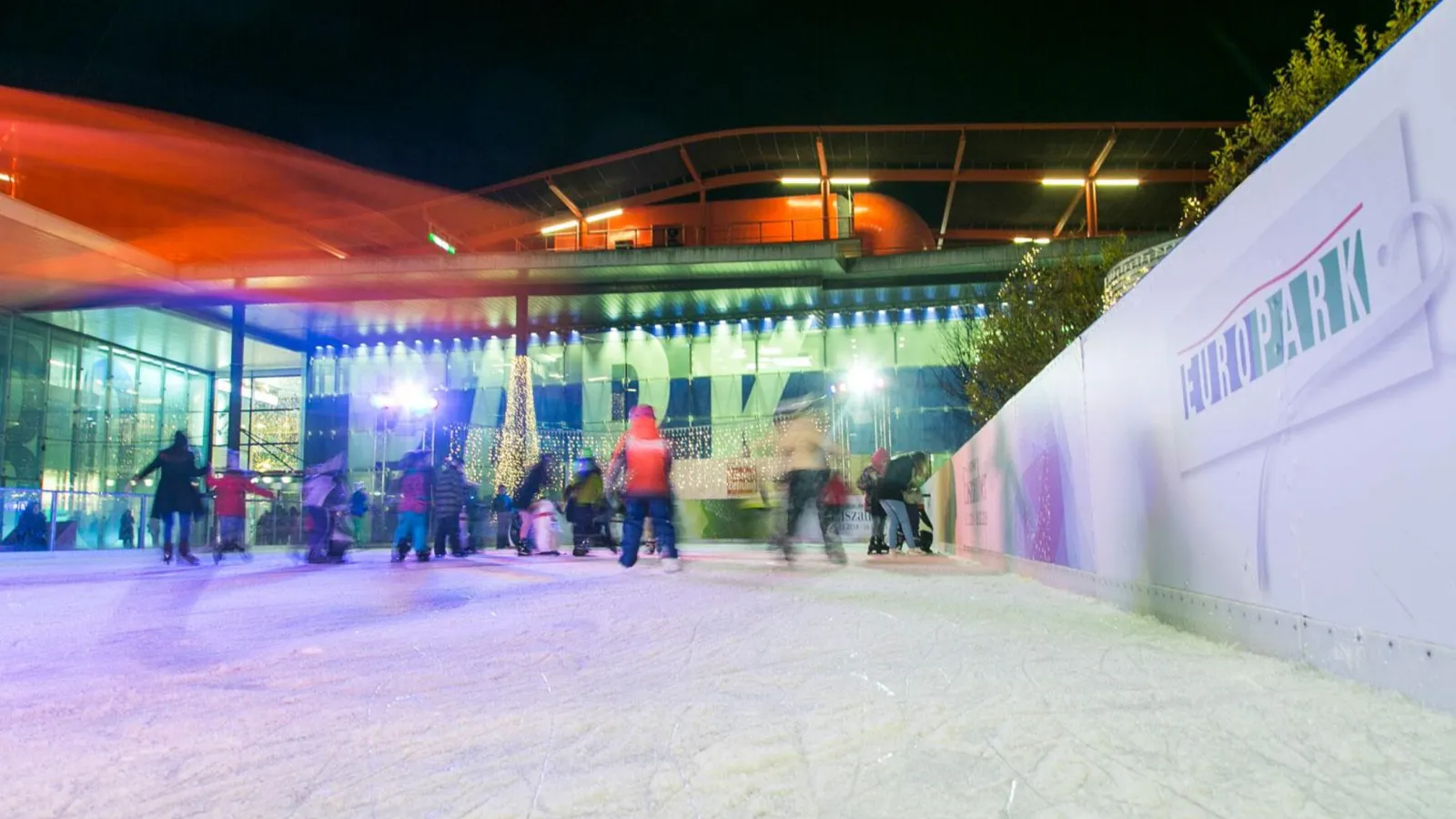 The Europark Eislaufplatz at night: skaters crossing the open ice inside the rink boards, with the shopping centre's lit entrance behind. — Gratis-Eislaufplatz Europark
