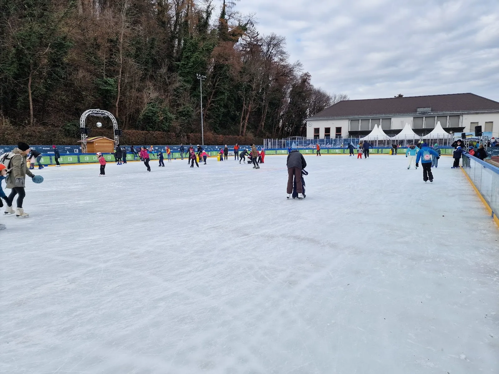 The Grazer Winterwelt open-air rink on a winter day, with skaters on the ice and the wooded hillside and the park building behind. — Grazer Winterwelt