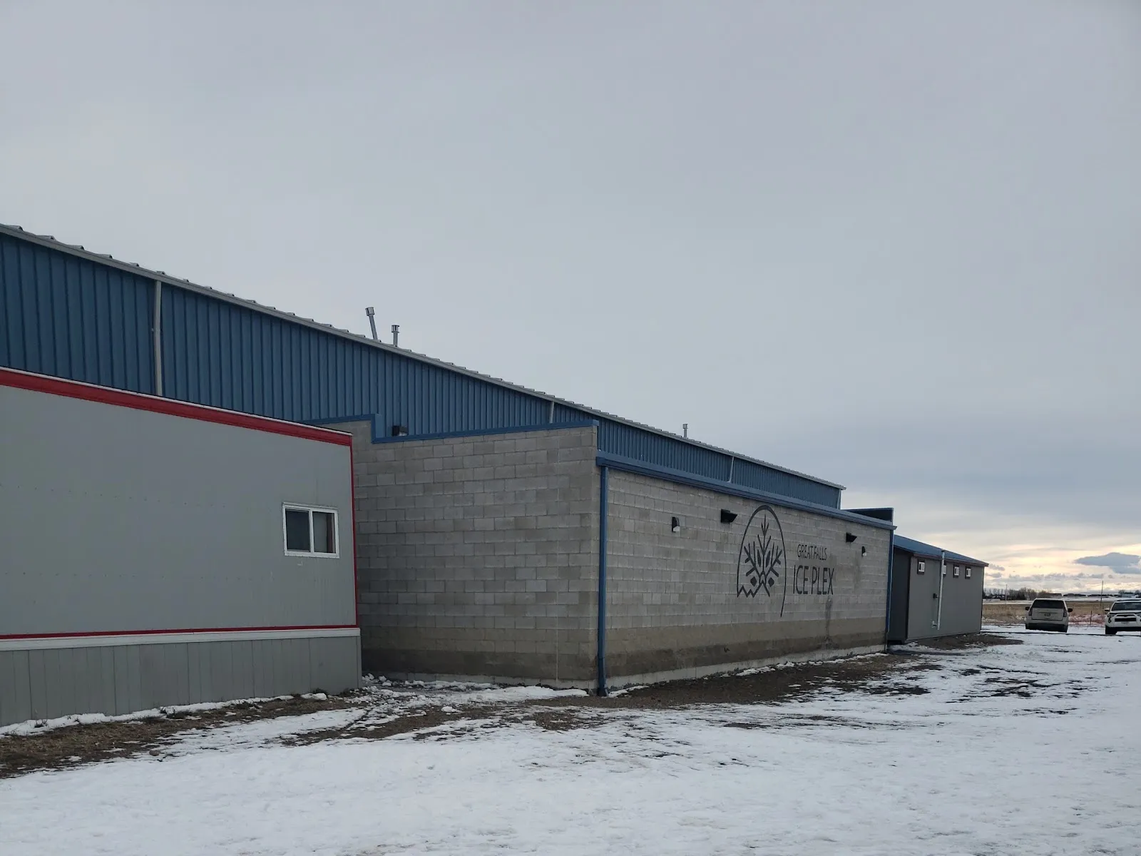 The Ice Plex's blue-and-gray metal building from the parking lot in winter, with the "Great Falls Ice Plex" logo and name painted on the cinderblock wall — Great Falls Ice Plex