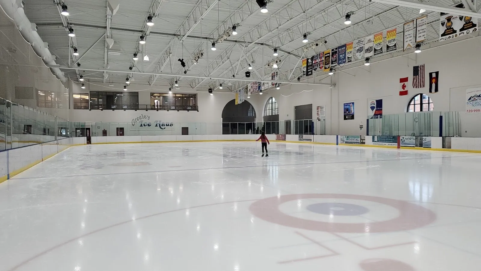 La pista de Greeley Ice Haus con un patinador en el hielo y su nombre en el muro del fondo, bajo ventanas en arco y mantas — Greeley Ice Haus