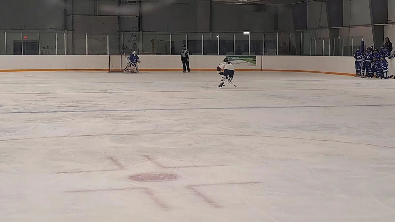 A wide view across the ice at Greenbriar Recreation Centre toward the far end, with a faceoff dot in the foreground, a goalie and an official by the net and a team on the bench. — Greenbriar Recreation Centre
