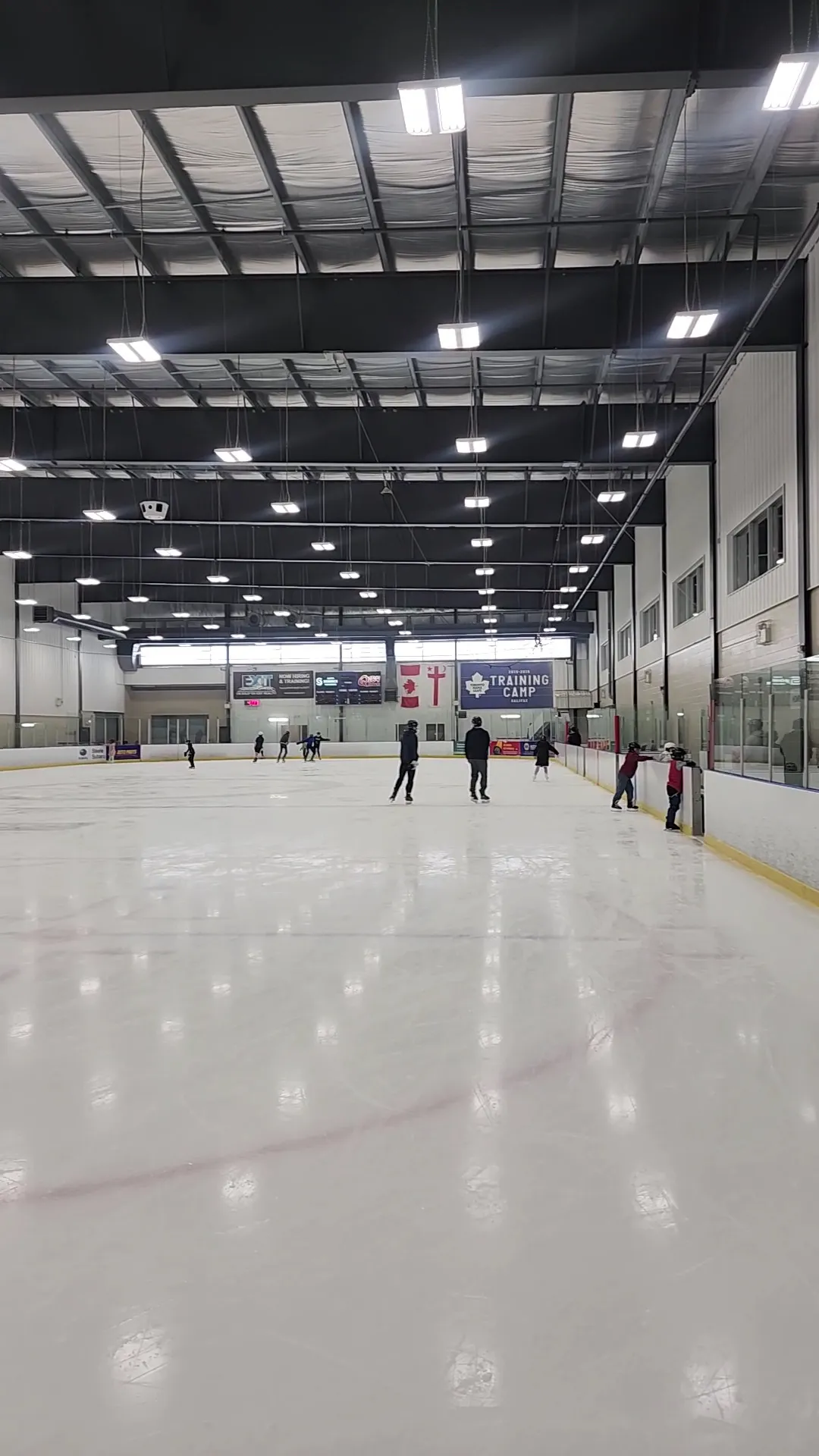 Skaters on the ice at the Greenfoot Energy Centre under the dark steel roof, with the sponsor banners and glass along the far side. — Greenfoot Energy Centre
