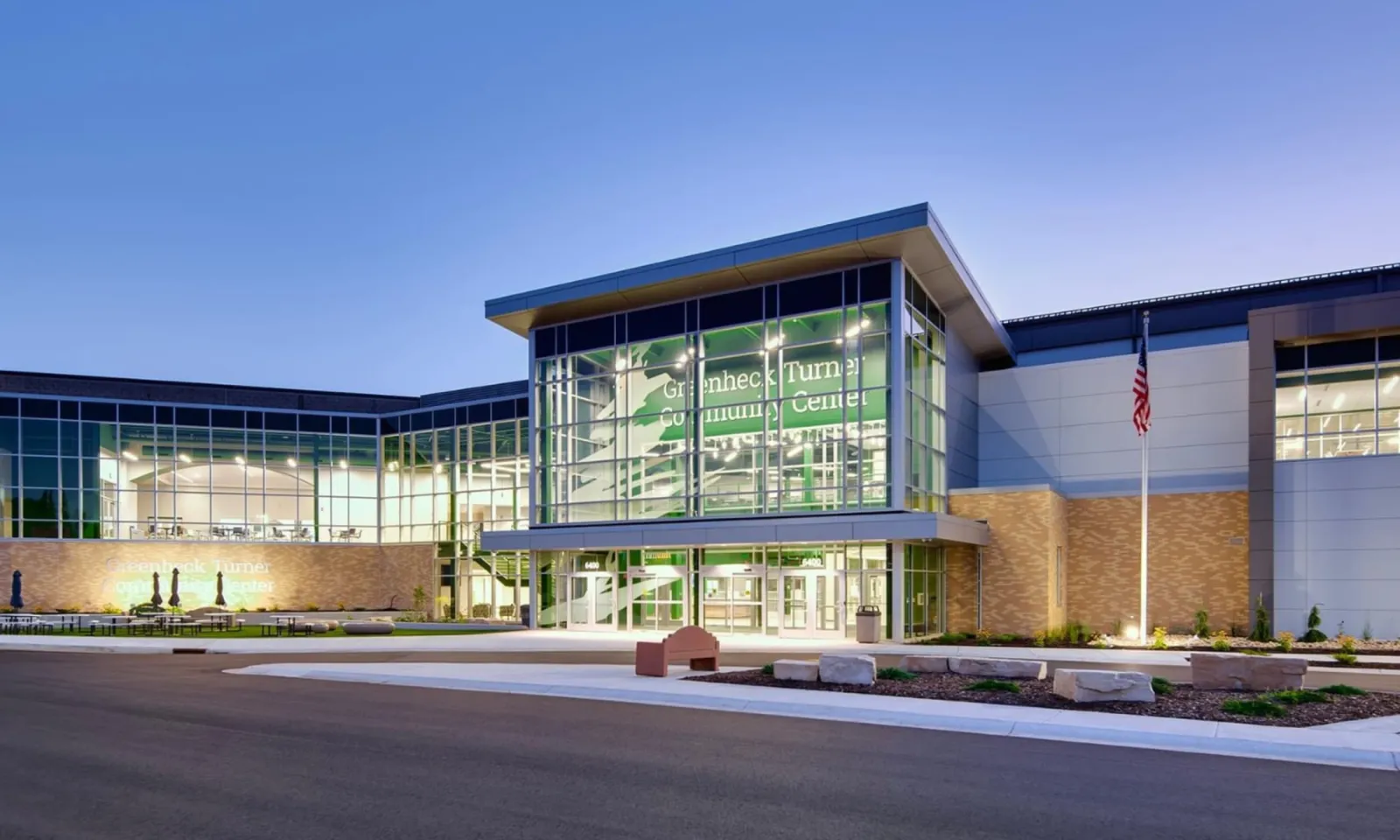Exterior al atardecer de la entrada del Greenheck Turner Community Center, con el atrio de vidrio iluminado con el nombre del edificio y una bandera de Estados Unidos al frente. — Greenheck Turner Community Center Ice Arena