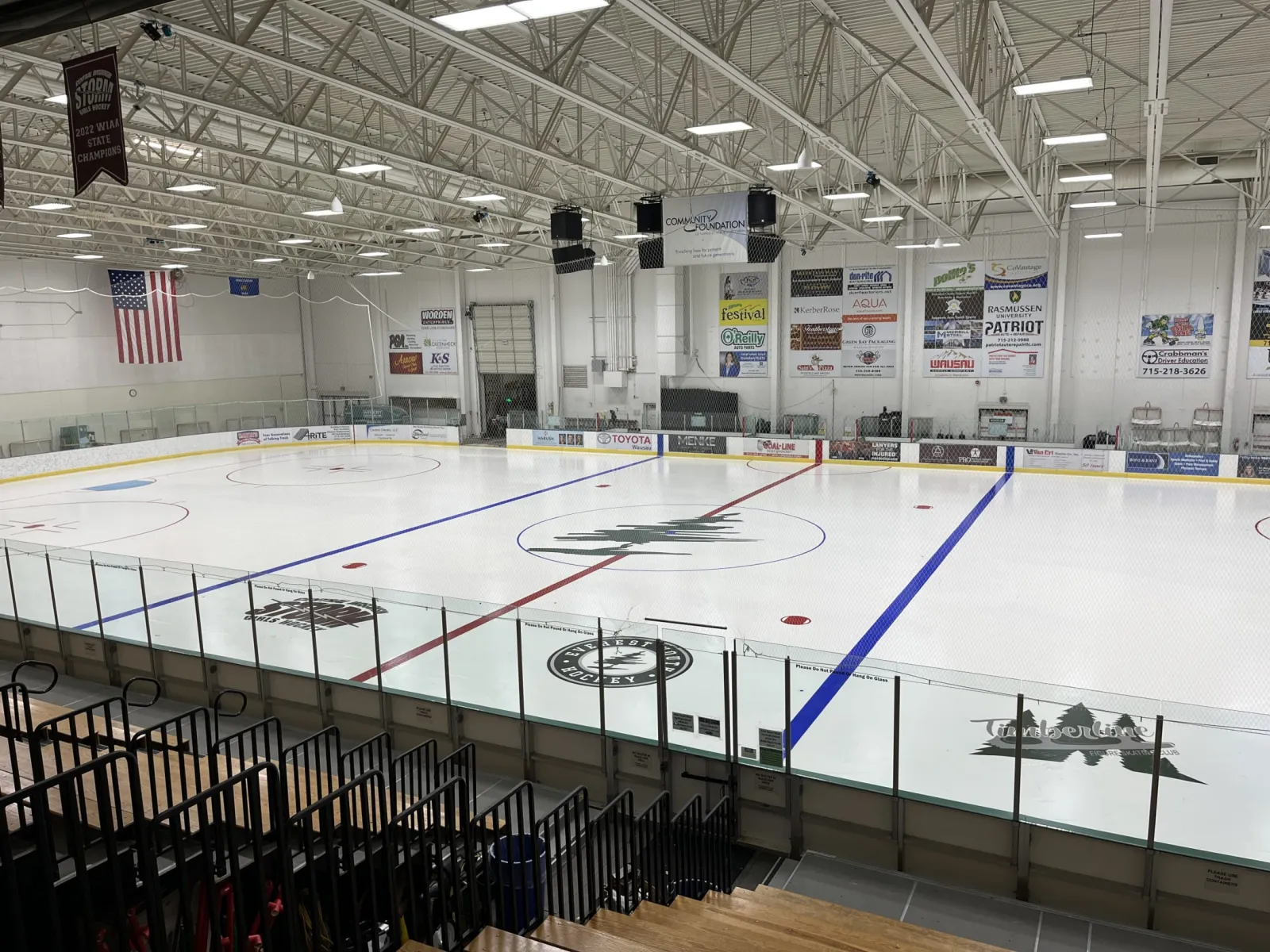 Pista vacía recién pulida con el logotipo central y las marcas de la pista, calcomanías de Everest Youth Hockey y del Timberline Figure Skating Club en las vallas y mantas de patrocinadores en las paredes. — Greenheck Turner Community Center Ice Arena