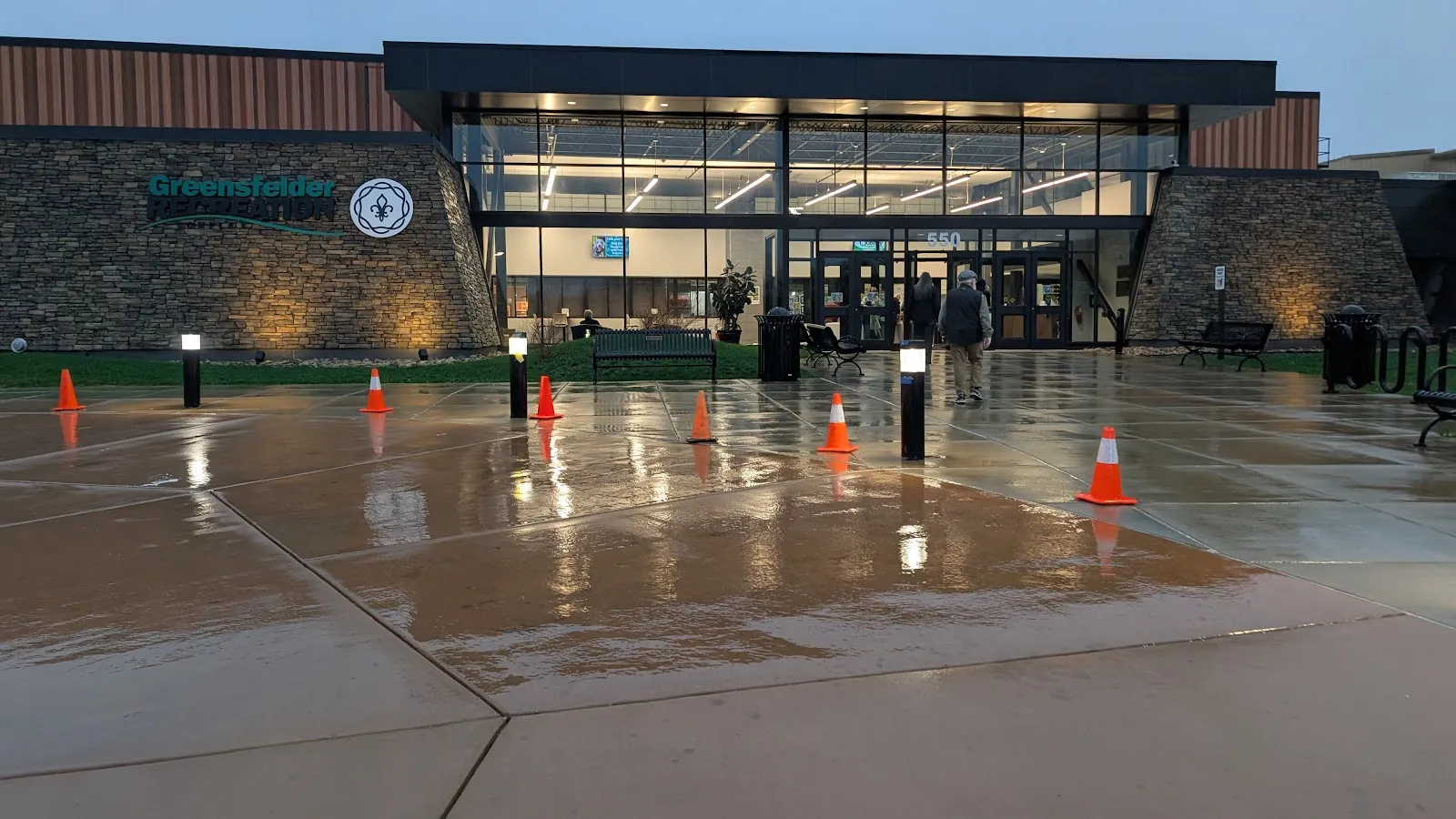 The glass-fronted entrance of the Greensfelder Recreation Complex at dusk, number 550 over the doors, stone wall with the complex name on the left and a wet paved forecourt in front. — Greensfelder Recreation Complex Ice Rink