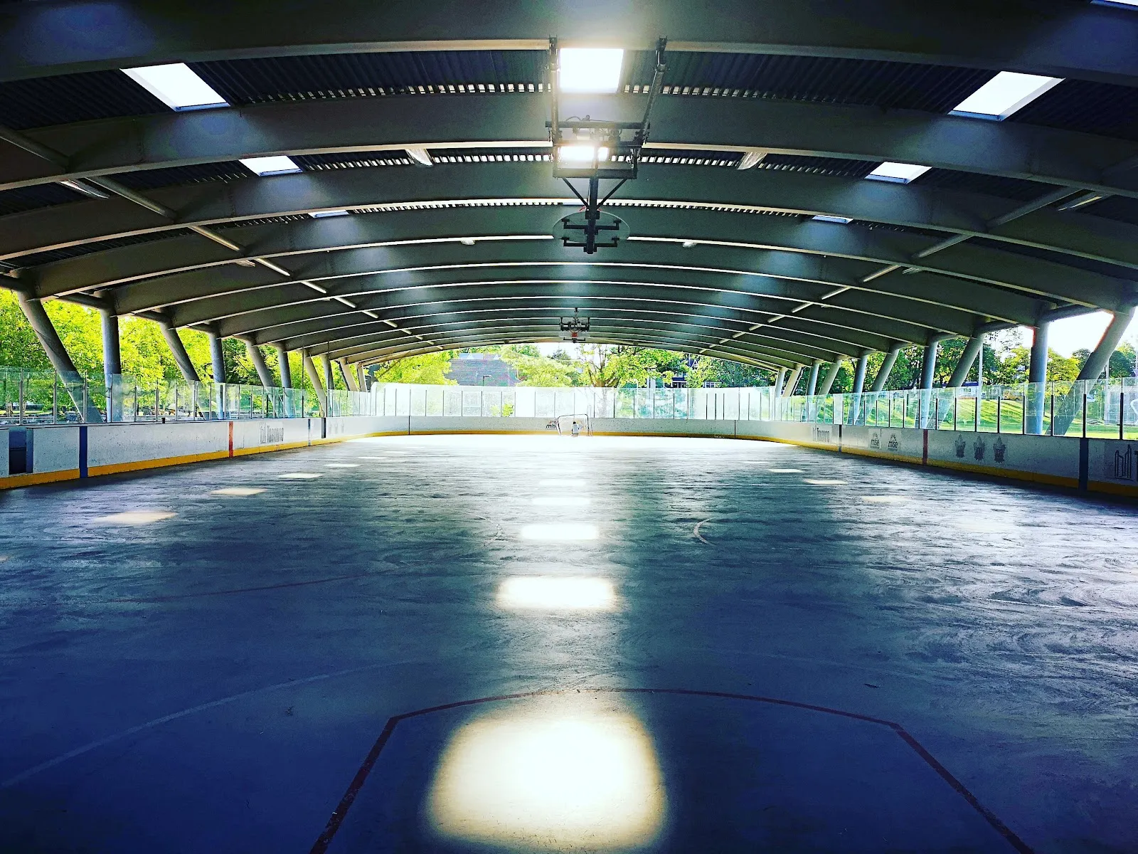 La pista techada de Greenwood vista a lo largo en la temporada cálida, con la superficie descubierta, bardas y vidrios, los reflectores reflejados en el piso y sin personas a la vista — Greenwood Park Outdoor Ice Rink and Skate Trail