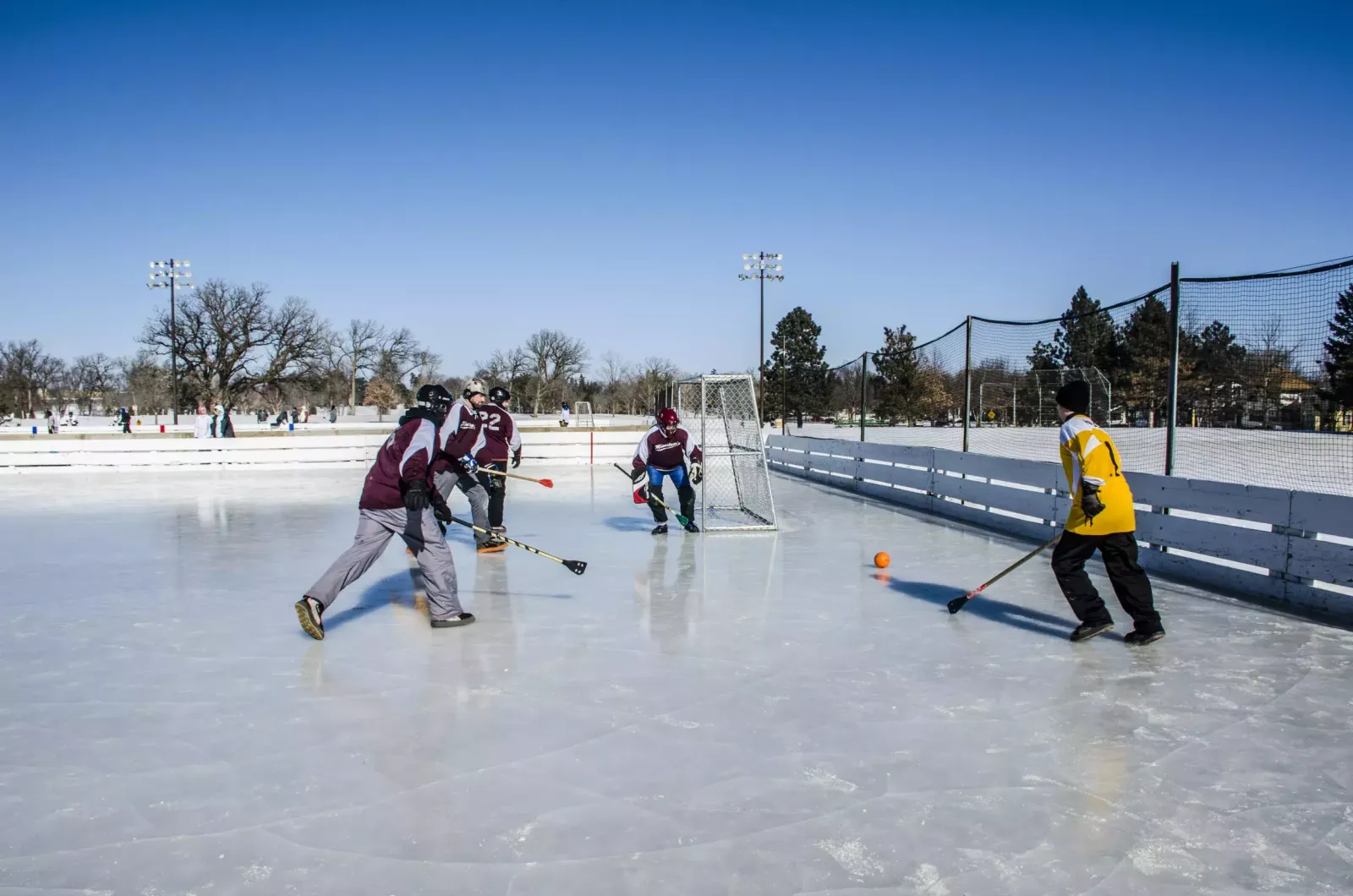 Players with sticks chase an orange ball toward a goal on the pale ice of the Groveland rink under a clear blue sky, with floodlight poles and a netted fence behind. — Groveland Park Outdoor Rinks
