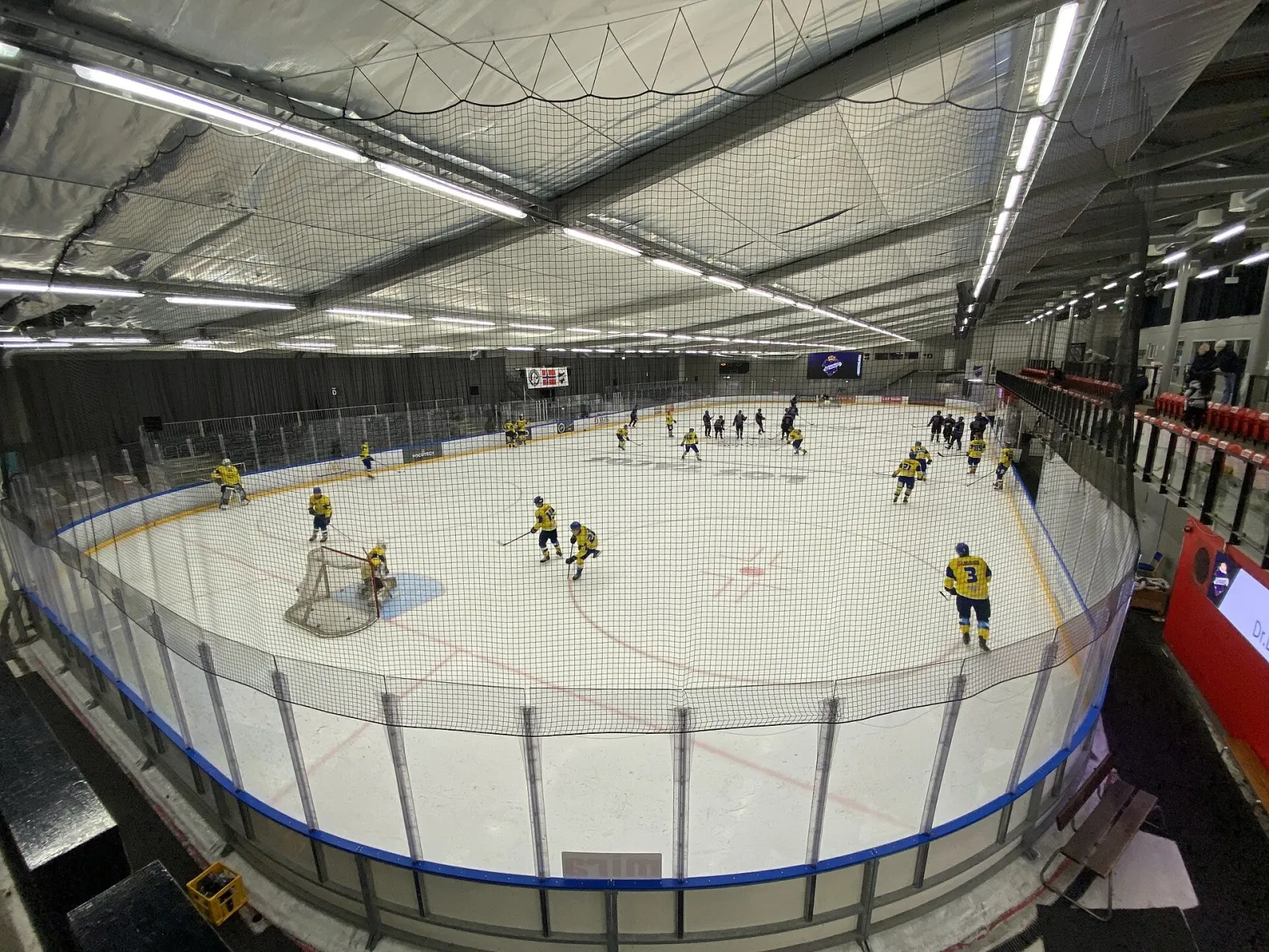 An ice hockey game in play in Grünerhallen, seen from above the boards with players spread across the ice and the roof trusses overhead. — Grünerhallen
