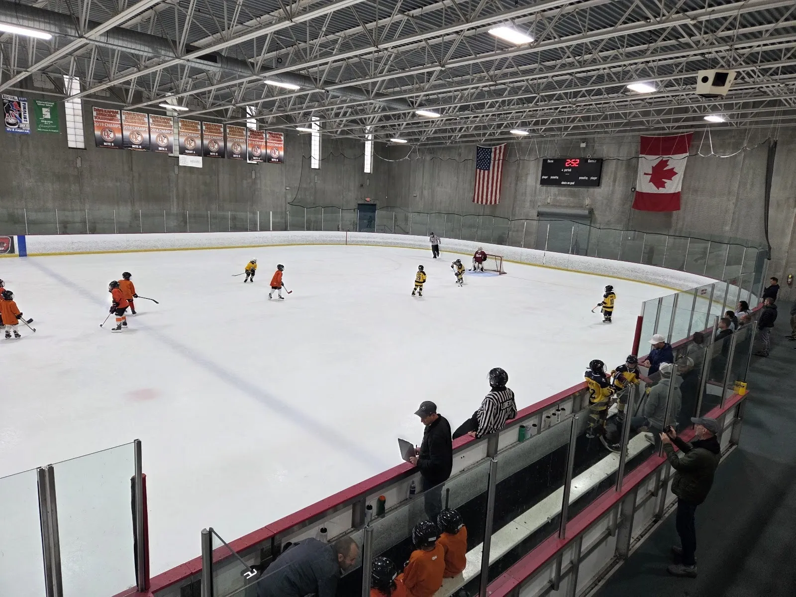 Un partido en un extremo de una pista amplia visto desde el vidrio, con jugadores repartidos por el hielo y banderas de Canadá y Estados Unidos y banderines en la pared del fondo. — Grundy Ice Arena