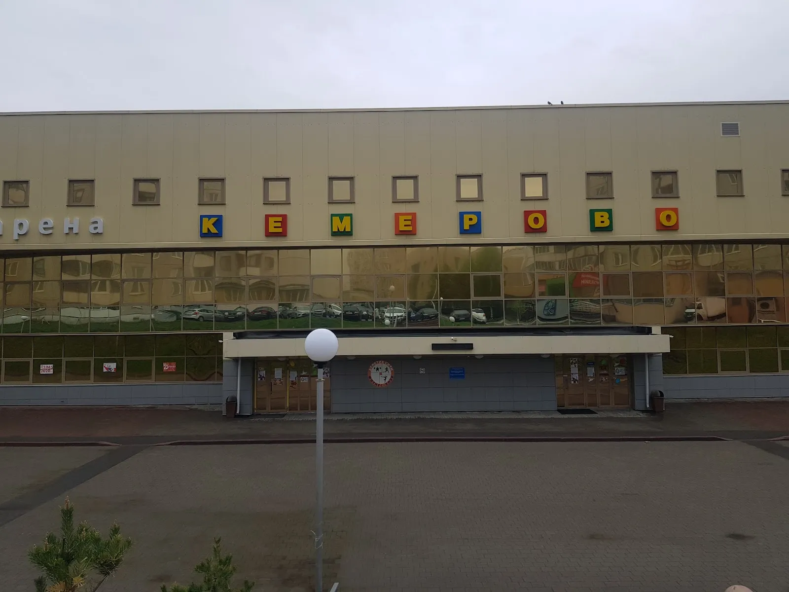 The front of the arena building on Molodezhny Prospekt, a beige panelled facade with gold-tinted glazing, coloured letters reading Kemerovo and the entrance canopy over a paved forecourt. — Gruzdev Ice Arena Kemerovo