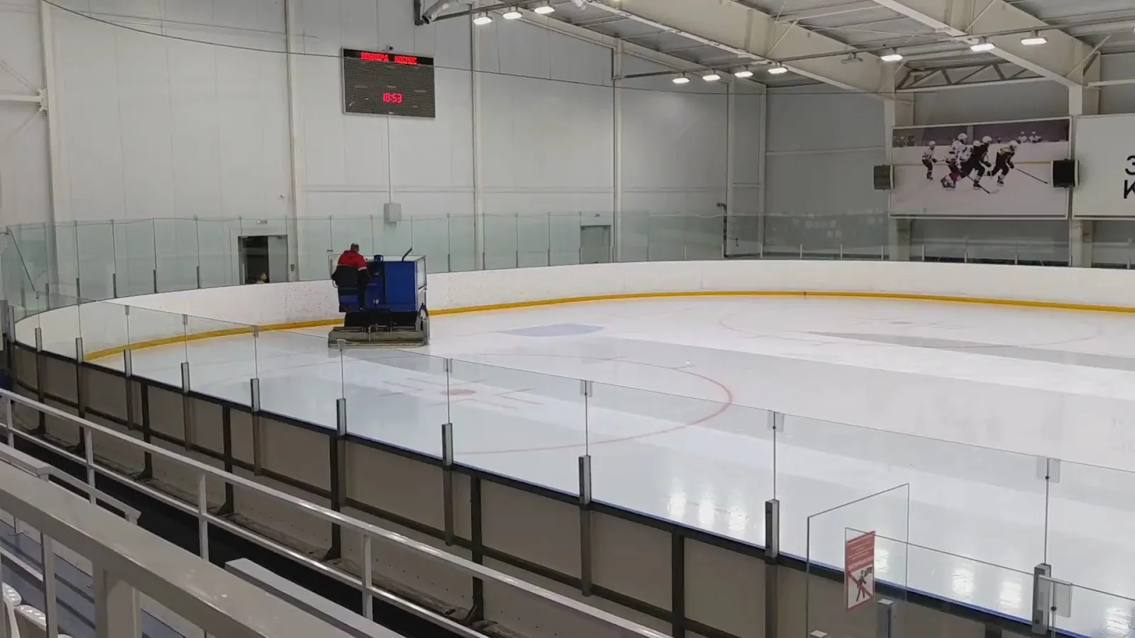 The indoor ice sheet seen from the stands, with glass above the boards, an ice resurfacer on the far side, a scoreboard and a banner of hockey players on the end wall. — Gruzdev Ice Arena Kemerovo