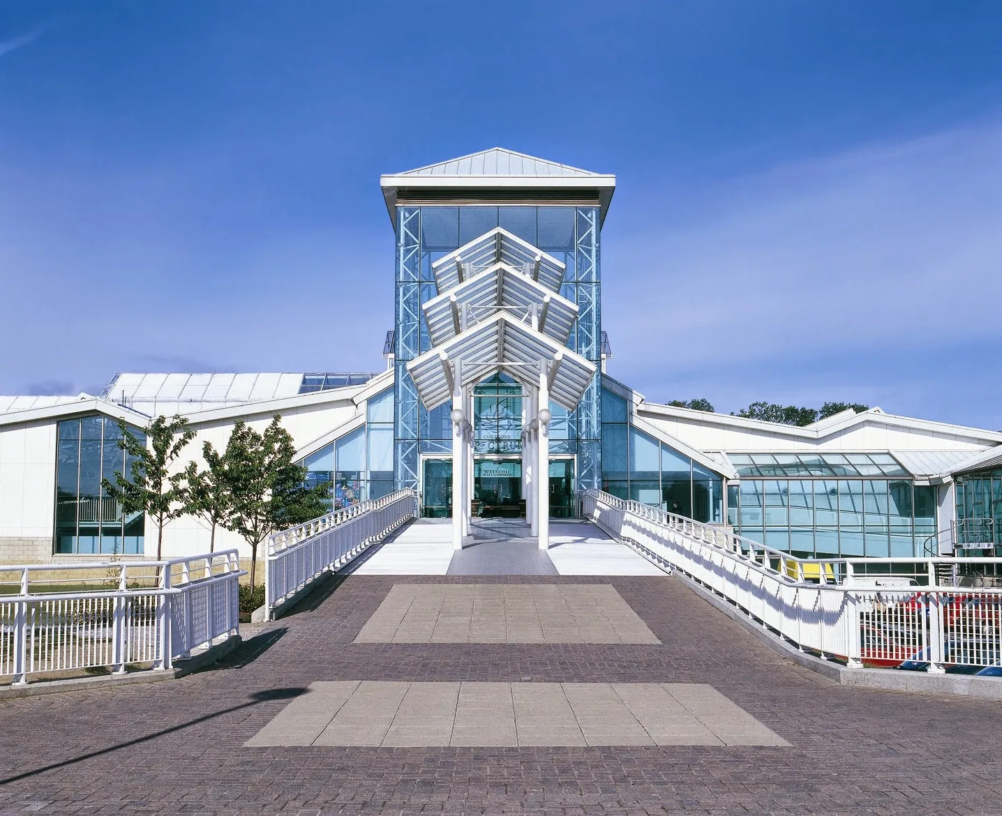 The glass and white-steel entrance tower of Guildford Spectrum at the top of a paved ramp, with the glazed leisure-centre roofs either side. — Guildford Spectrum