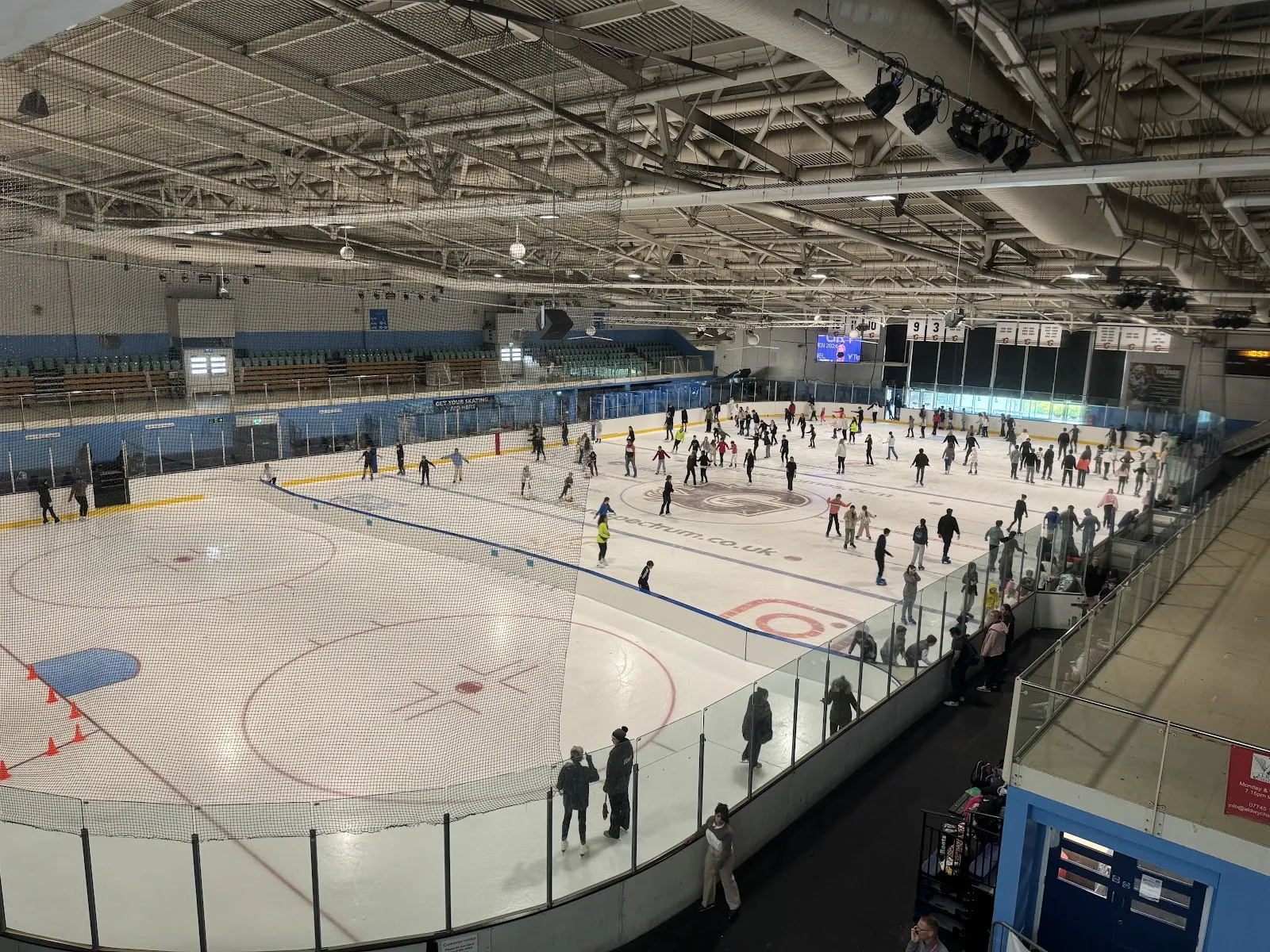 The ice pad from the raised viewing walkway during a public session, with many skaters on the far half of the sheet and the steel roof trusses above. — Guildford Spectrum
