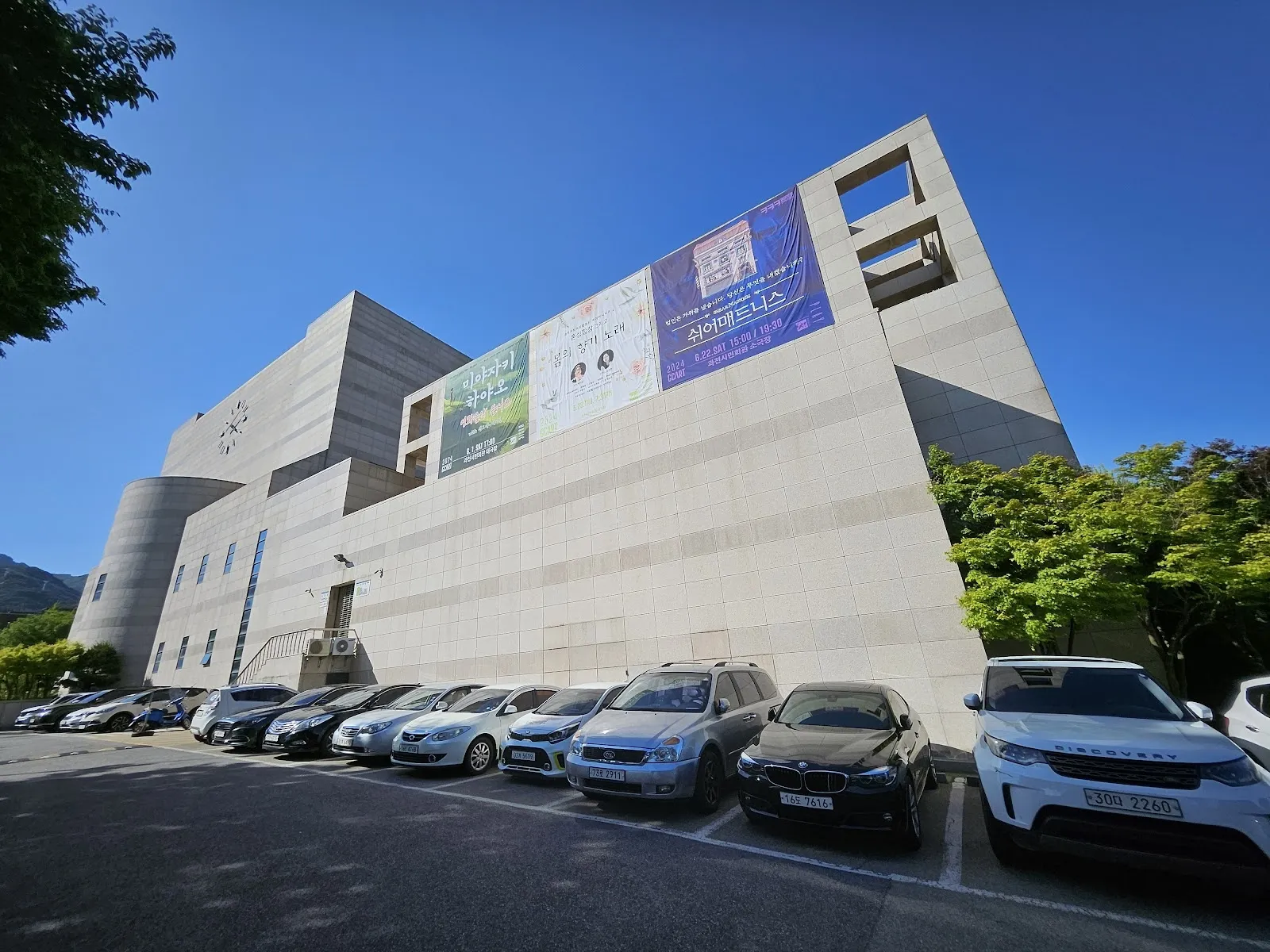 아이스링크가 들어 있는 과천시민회관의 회색 콘크리트 외벽과 건물을 따라 주차된 차량들. — Gwacheon Civic Hall Ice Rink