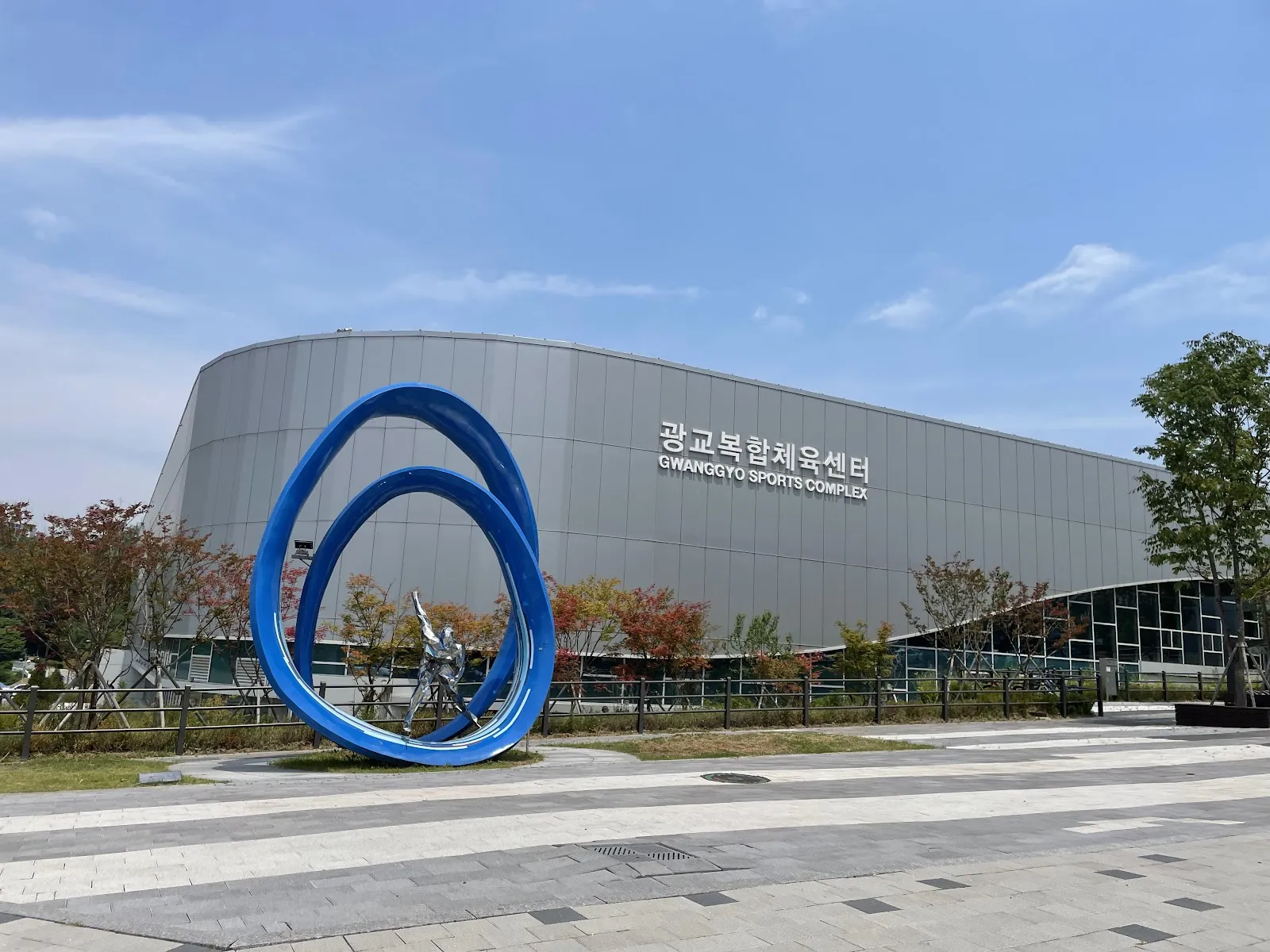 The grey curved facade of Gwanggyo Complex Sports Center with its name in steel letters and a blue loop sculpture in the plaza. — Gwanggyo Ice Rink (Suwon)