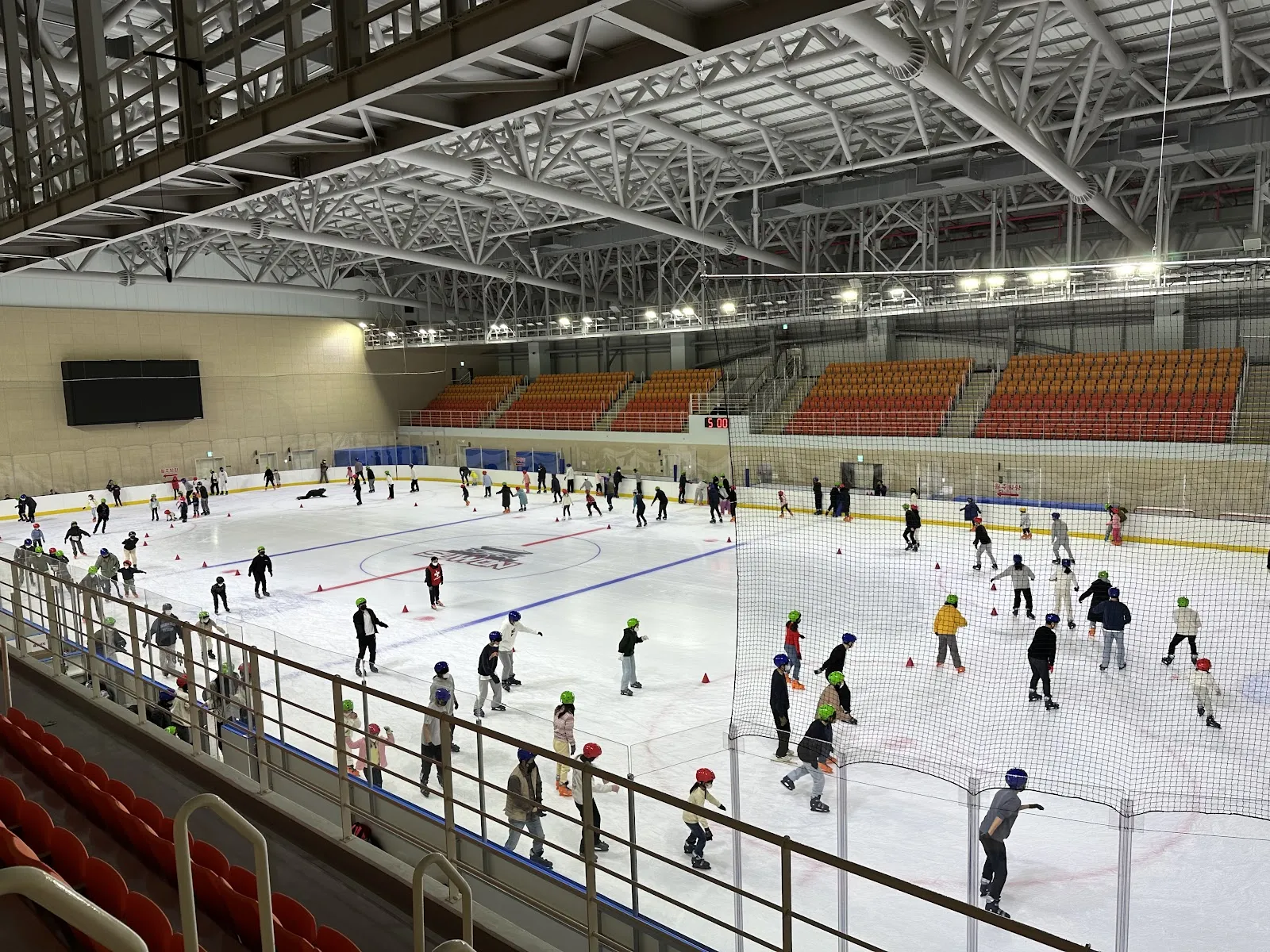 The ice rink of Gwanggyo Complex Sports Center from the stand during a public session, a wide view of the lit hall with the orange seating and skaters spread over the ice. — Gwanggyo Ice Rink (Suwon)