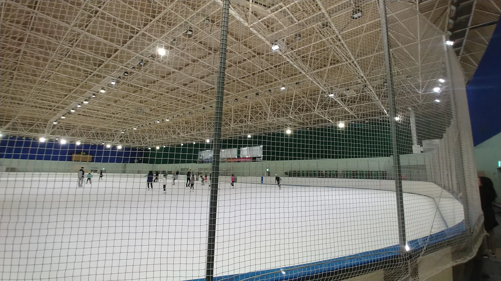 안전망 뒤로 보이는 광주 실내빙상장의 빙판. 철골 트러스 구조의 경기장이 넓게 보이고 멀리 스케이터 몇 명이 있어요. — Gwangju Indoor Ice Rink