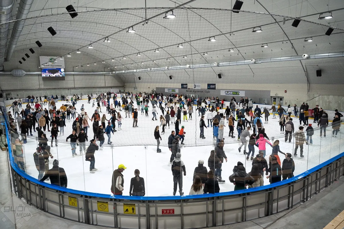 A crowded public skating session on the ice of the Győr university ice sports centre, with skaters across the whole sheet and the arched roof above. — Győri Egyetemi Jégsport Központ