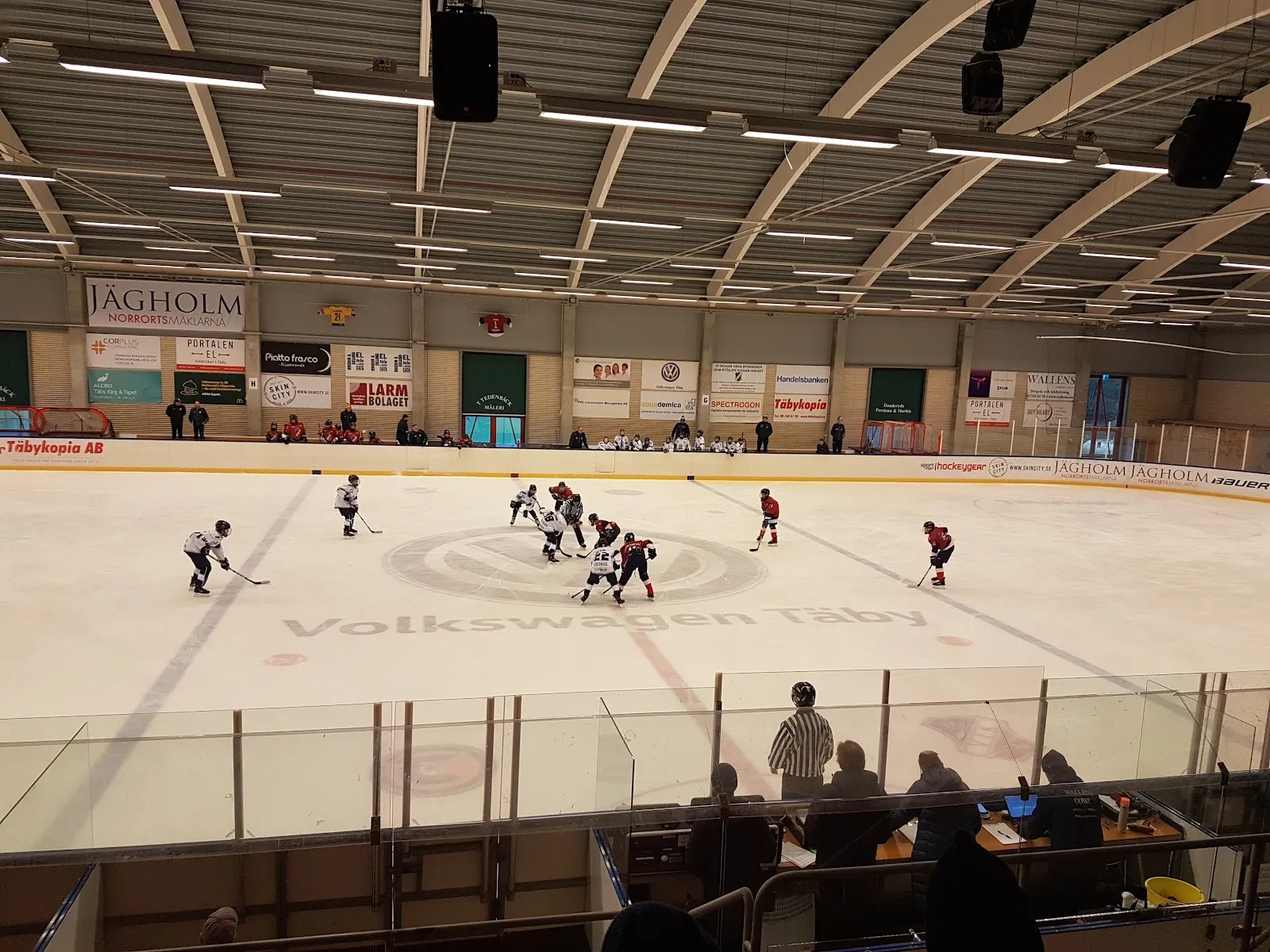 Youth hockey game on the ice sheet in Hägernäs ishall (Jägholmshallen), seen from above the benches with the sponsor boards behind – Hägernäs ishall