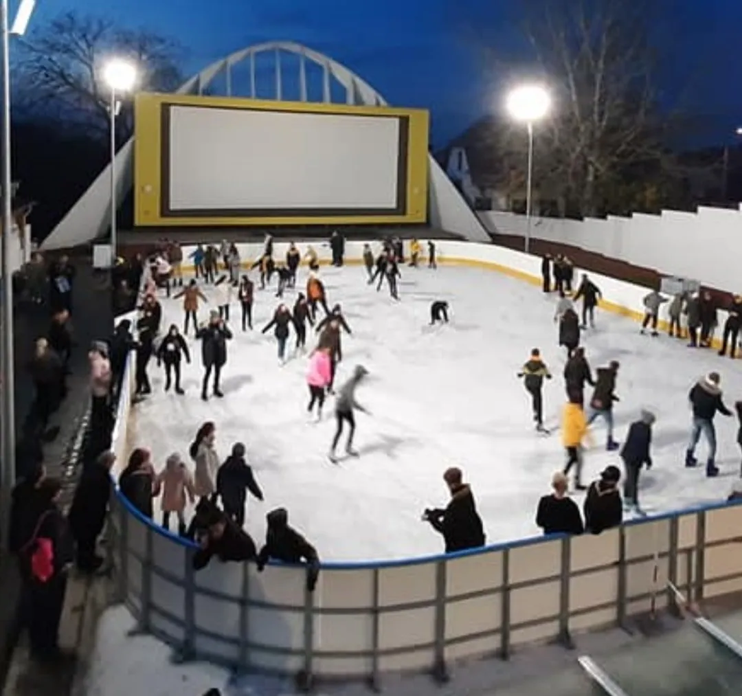 A public skating evening on the open-air rink in Hajdúszoboszló, with skaters spread across the white ice under floodlights and a large screen wall behind an arched structure. — Hajdúszoboszlói Jégpálya