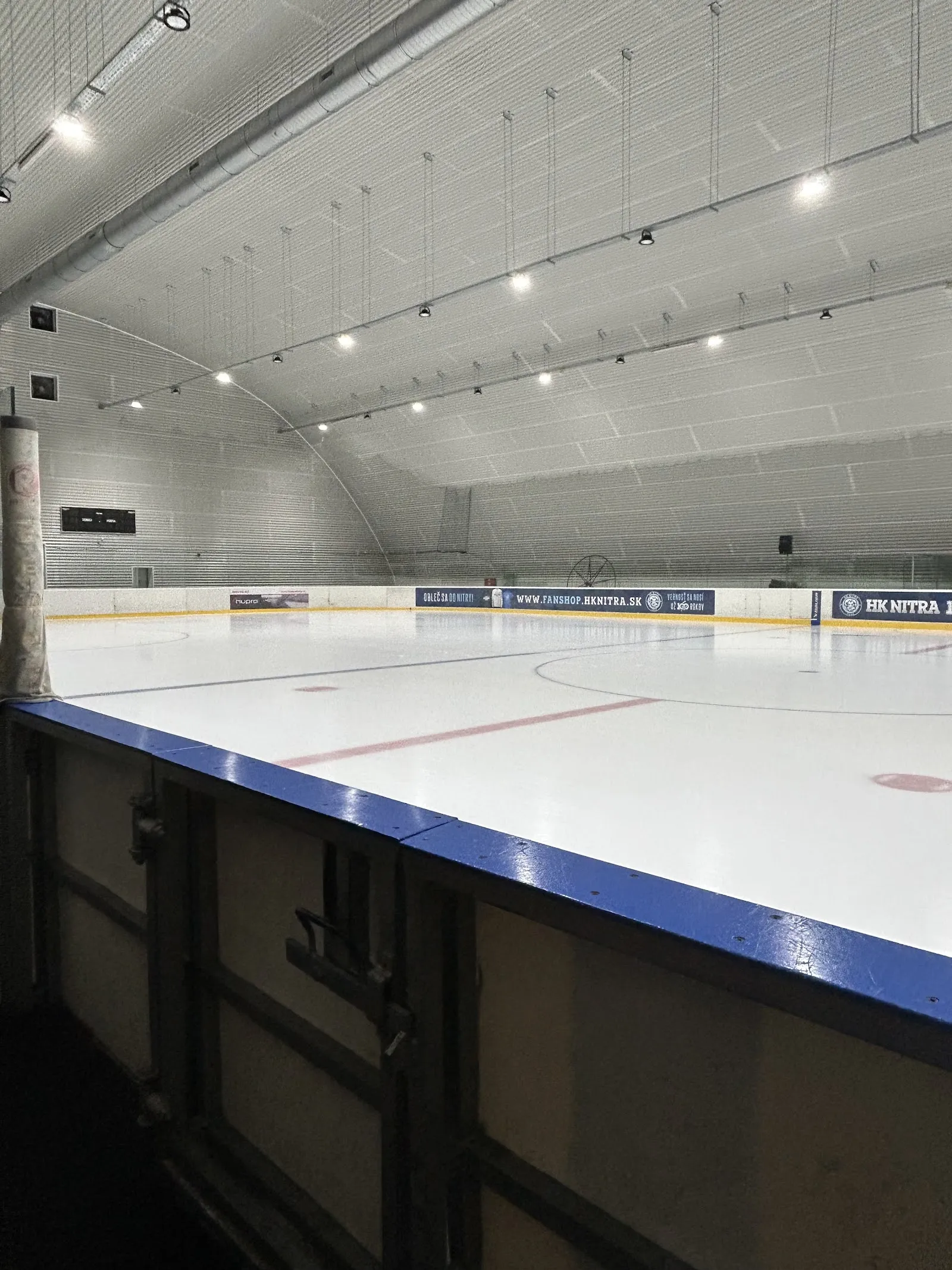 The empty ice sheet of Hala Klokočina seen from the barrier, with the HK Nitra boards along the far side and the curved hall roof above. — Hala Klokočina (HK Nitra)