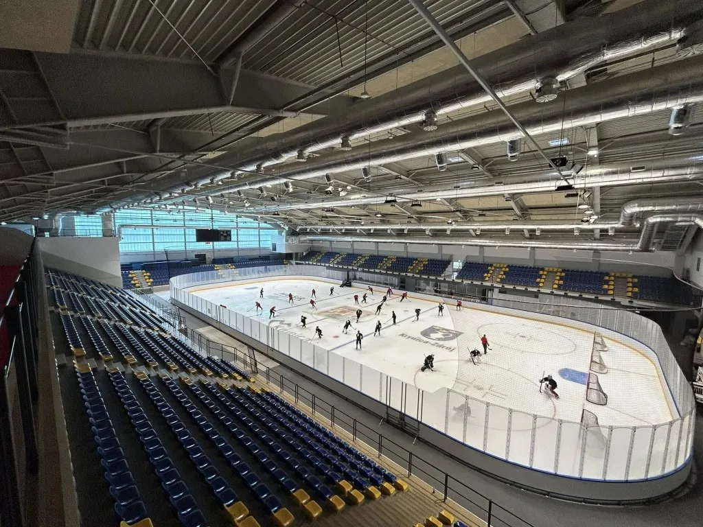 The ice of the Hala Lodowa MOSiR in Krynica-Zdrój seen from the high stands, with a few skaters far off on the full-size sheet and rows of blue seats around it — Hala Lodowa MOSiR Krynica-Zdrój