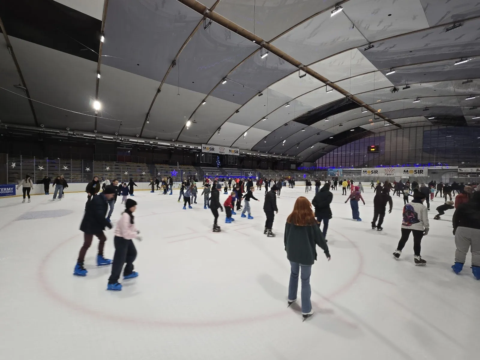 A public skating session on the ice of the Zgierz ice hall, with skaters across the sheet under the grey hall roof. — Hala Lodowa MOSiR Zgierz