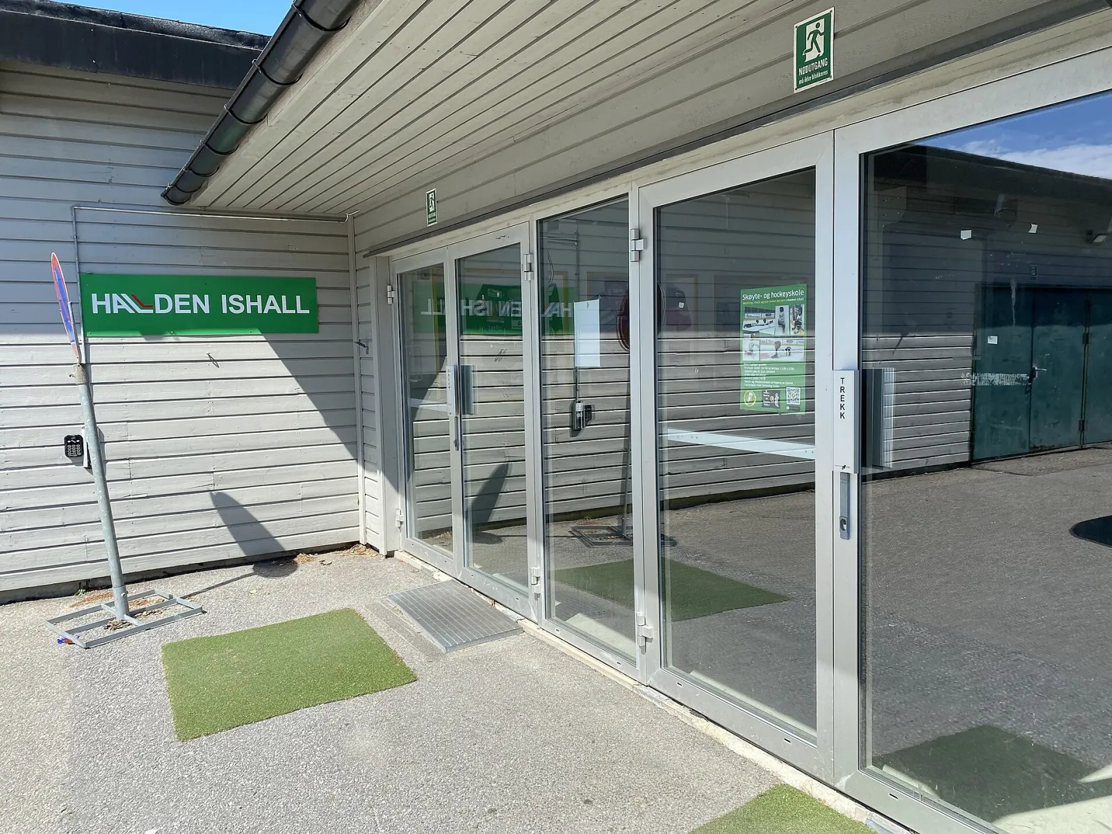 The glass entrance doors of Halden Ishall under the green name sign on the grey timber wall. — Halden Ishall