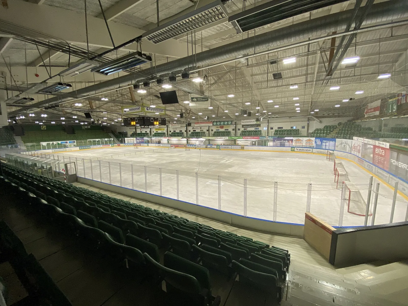 The ice sheet inside Halden Ishall seen from the green seats of the stand, with the boards and sponsor panels around the rink. — Halden Ishall