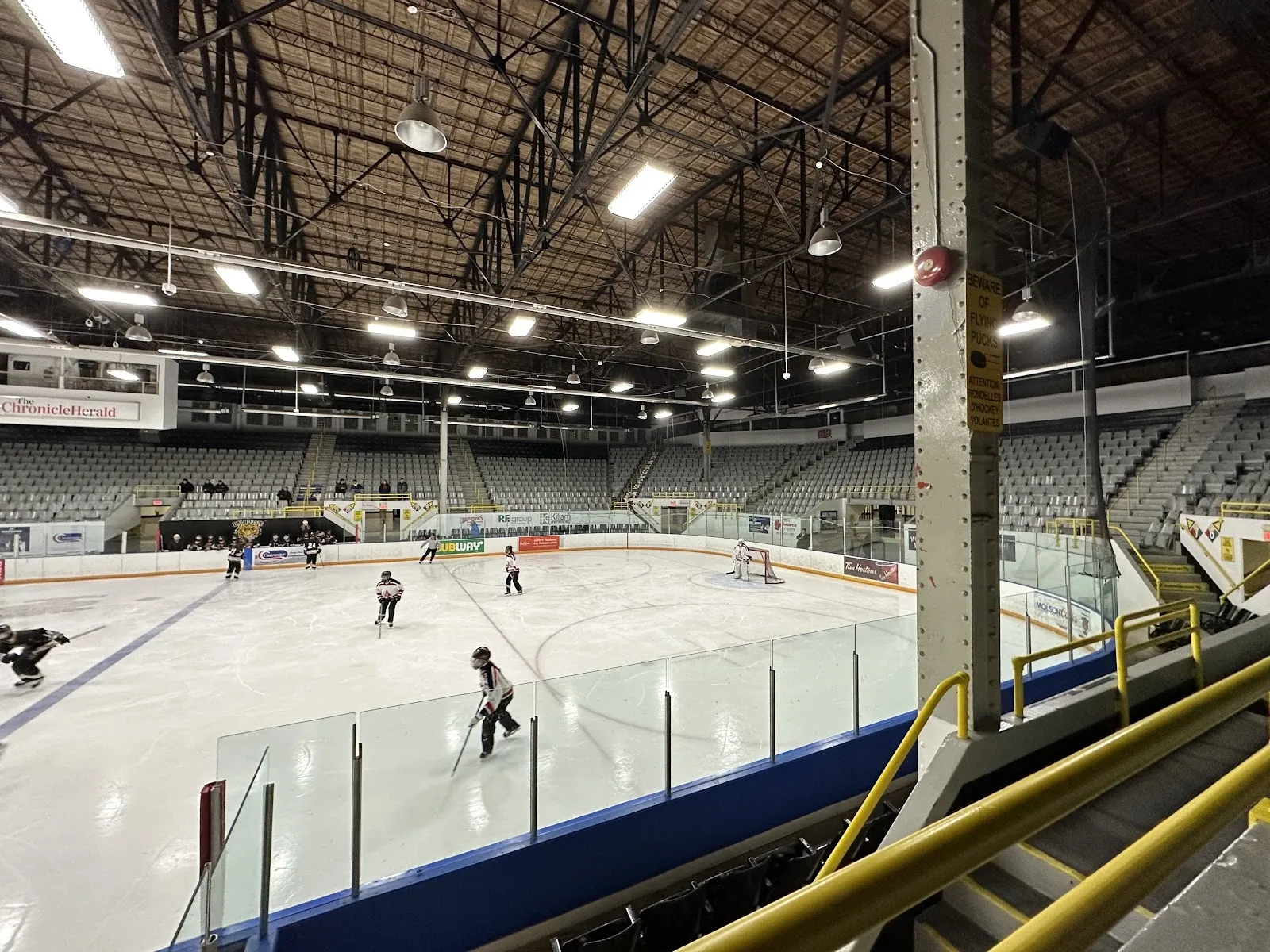 A game seen from the stands, players spread across a wide sheet with long blue-edged seating along the near side and a dark roof overhead. — Halifax Forum