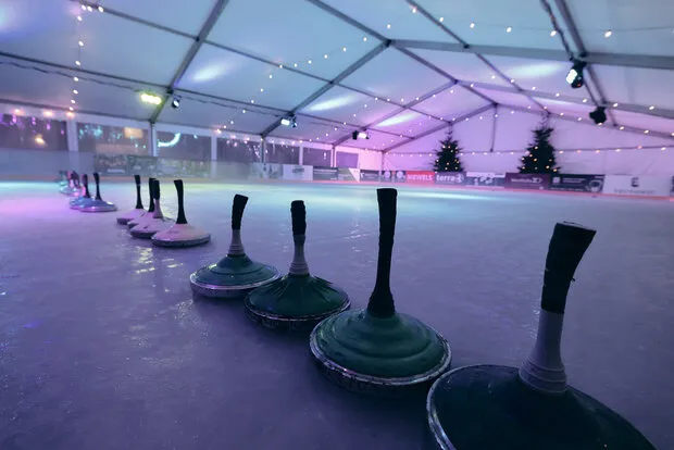 The ice rink of Halle on Ice under its tent, with a row of curling stones on the empty ice and strings of lights along the roof. — Halle on Ice