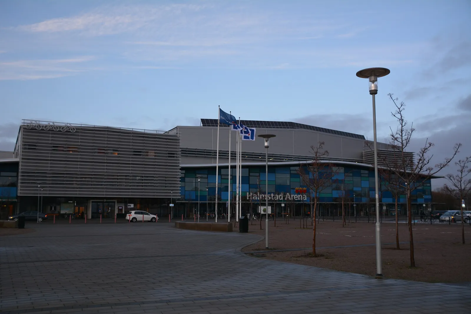 Framsidan av Halmstad Arena med glasfasad och flaggor, sedd över torget (2015) – Halmstad Arena (ishallarna)