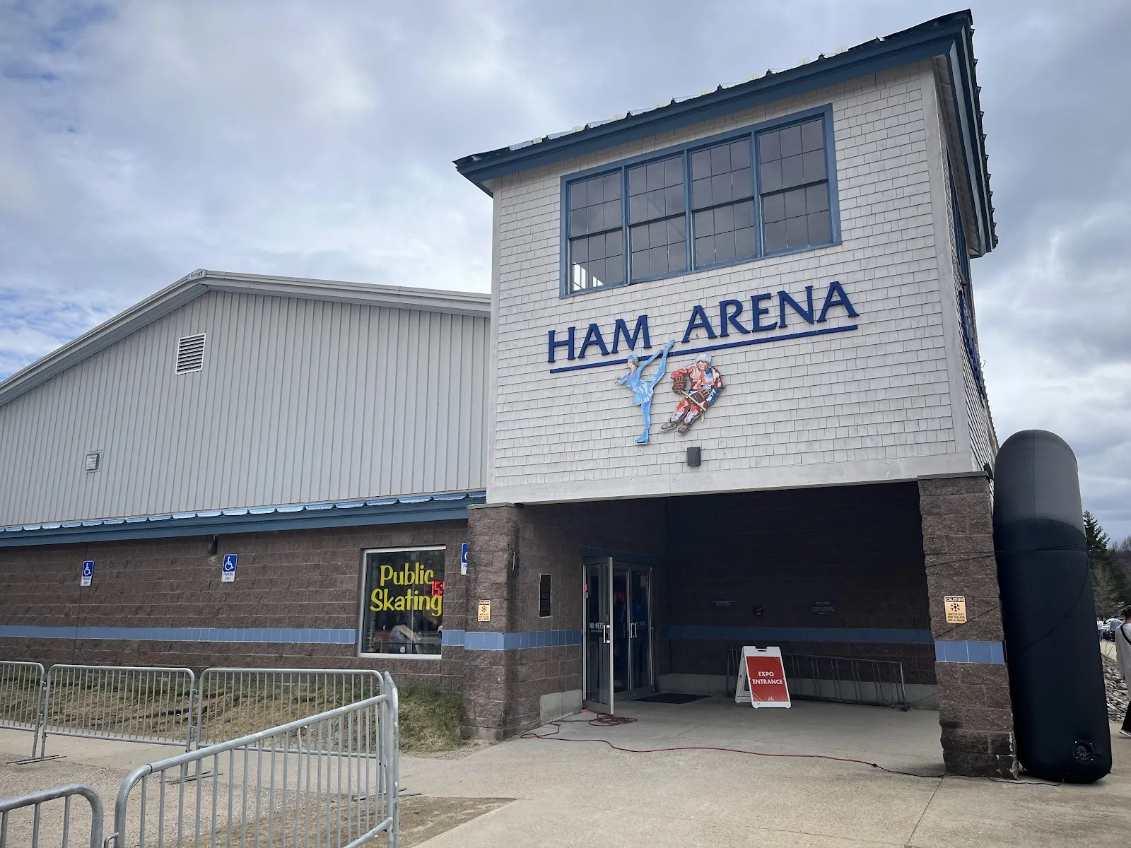 Front entrance tower of Ham Arena, white shingle siding with a large "HAM ARENA" sign and figure-skater/hockey-player cutouts above the doors, a "Public Skating" sign in the window, and the main gable-roofed rink building extending to the left. — Ham Arena