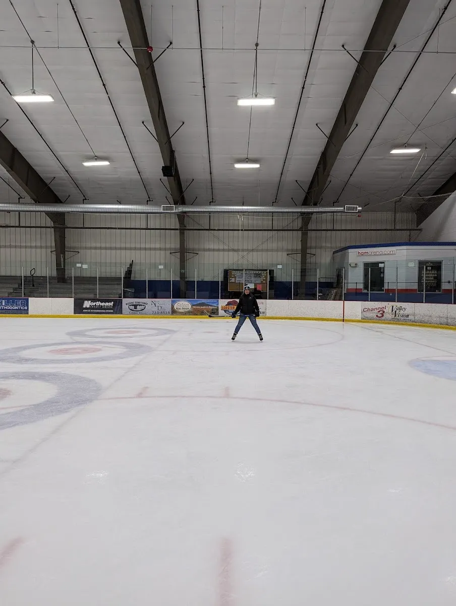 Most of the ice sheet, empty except for one adult skater near center ice, with curling-circle markings on the ice, dasher-board ads, bleachers, and the arena's own blue-and-white "hamarena.com" scorekeeper's booth visible along the back wall. — Ham Arena