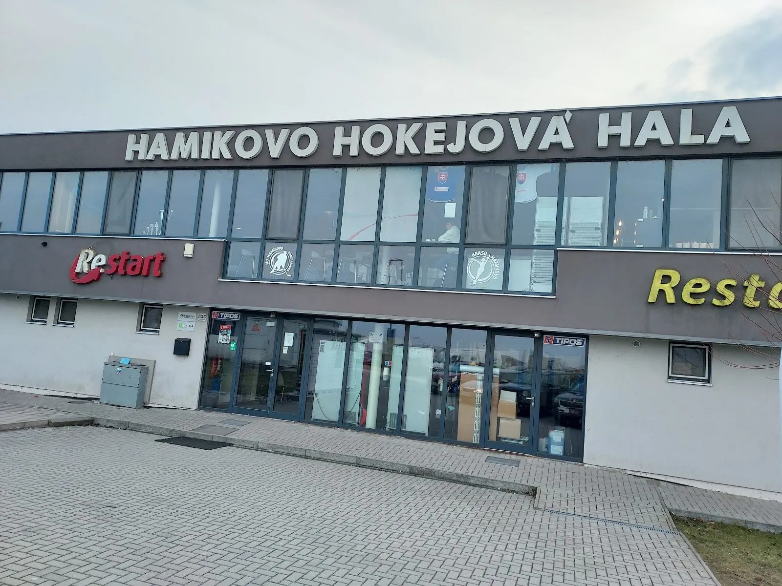 The glass-fronted Hamikovo hockey hall with its name along the top and a restaurant sign by the entrance. – Zimný štadión Hamikovo