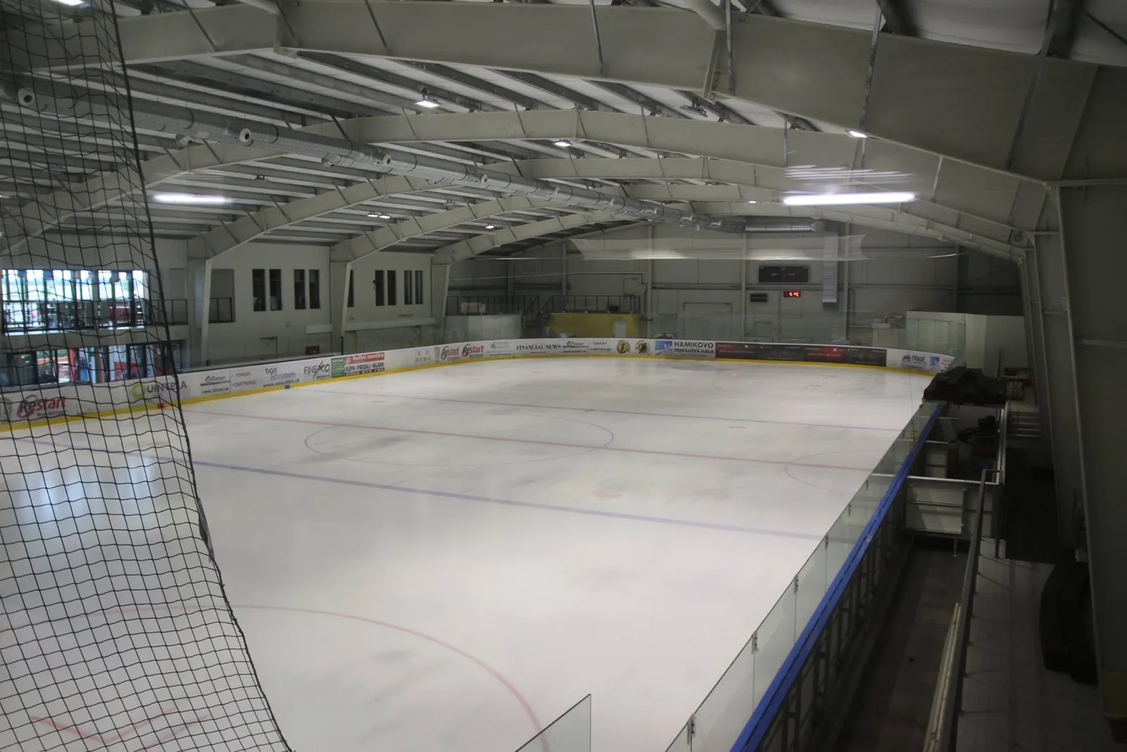 The empty ice sheet of the Hamikovo hall seen from the stands through the safety net, with boards, a steel-truss roof and the far-end glass wall. – Zimný štadión Hamikovo