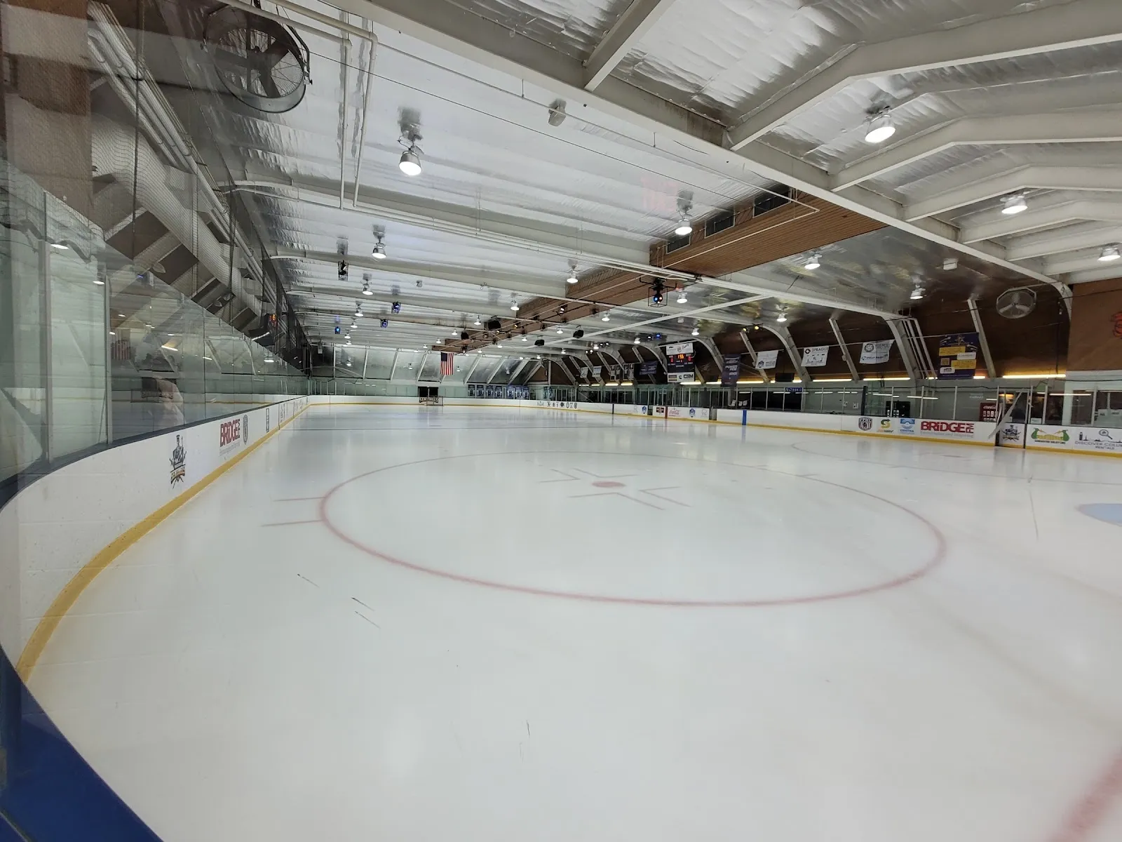 Una pista amplia y vacía a lo largo del vidrio, con el círculo central rojo sobre hielo blanco bajo un techo curvo de madera con banderines en lo alto de la pared del fondo. — Hamilton Center Ice Arena