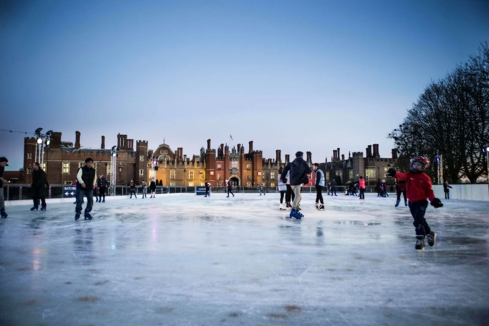 Skaters on the outdoor rink in front of Hampton Court Palace on a bright winter day, with the palace's brick facade behind the ice. — Hampton Court Palace Ice Rink
