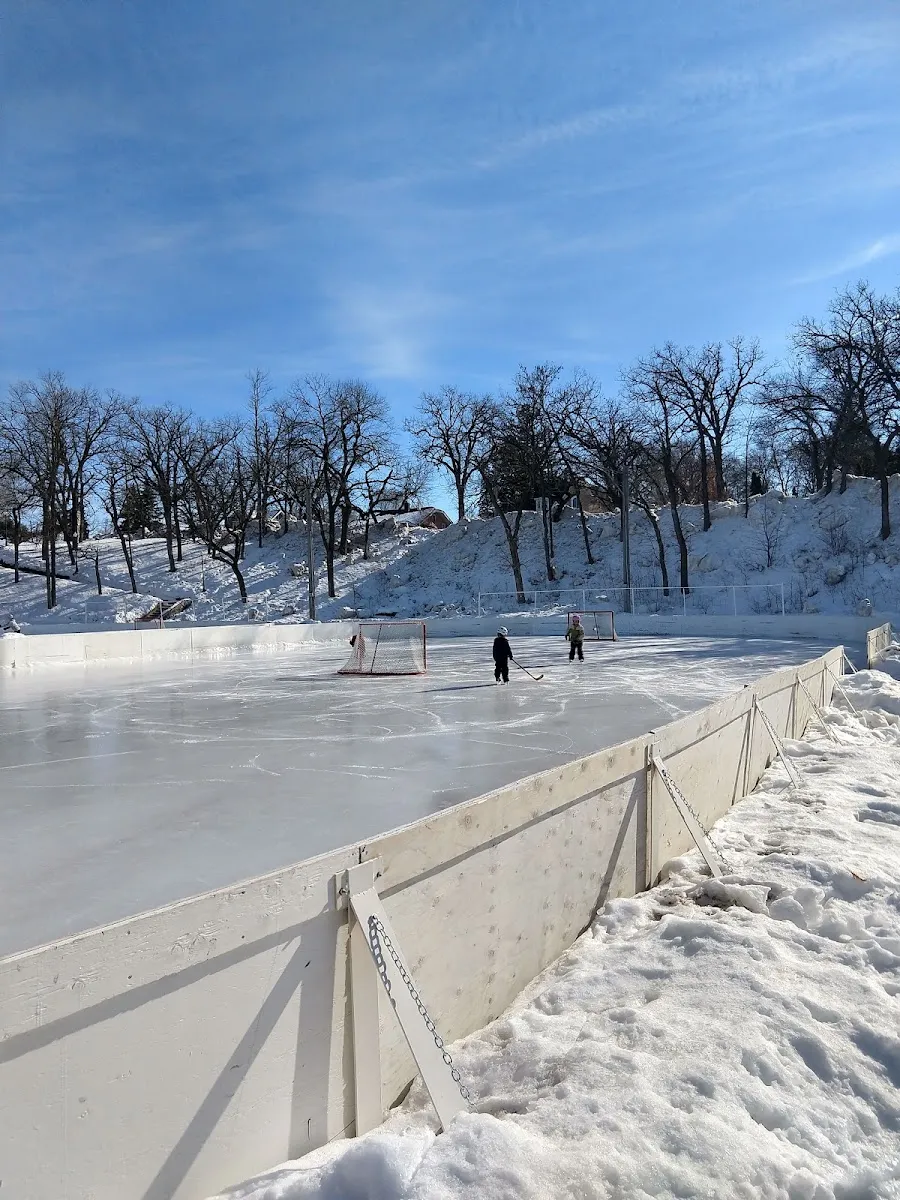 Two players at the far end of the board-enclosed rink at Handke Pit in winter, with trees on the bank above — Handke Pit Ice Rinks