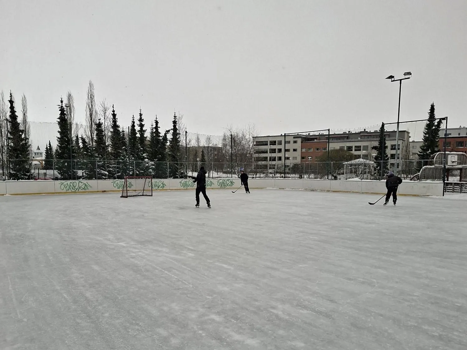 The boarded Hapelähde rink in winter with a few skaters and hockey players at a distance, a goal, floodlights and apartment blocks behind. — Hapelähteen jääkiekkokaukalo