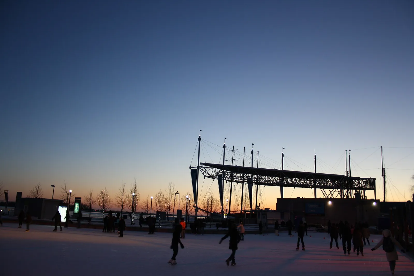 Skaters on the Harbourfront Centre rink at dusk beside the water, with the masts of a tall ship and the lit shoreline behind — Harbourfront Centre Skate by the Lake Rink
