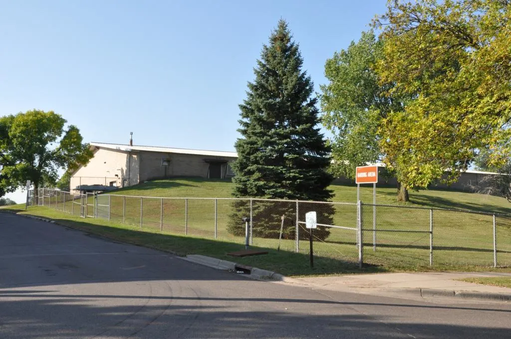 The low arena building behind a fence, lawn and a tall spruce, seen from the street on a clear day. — Harding Arena