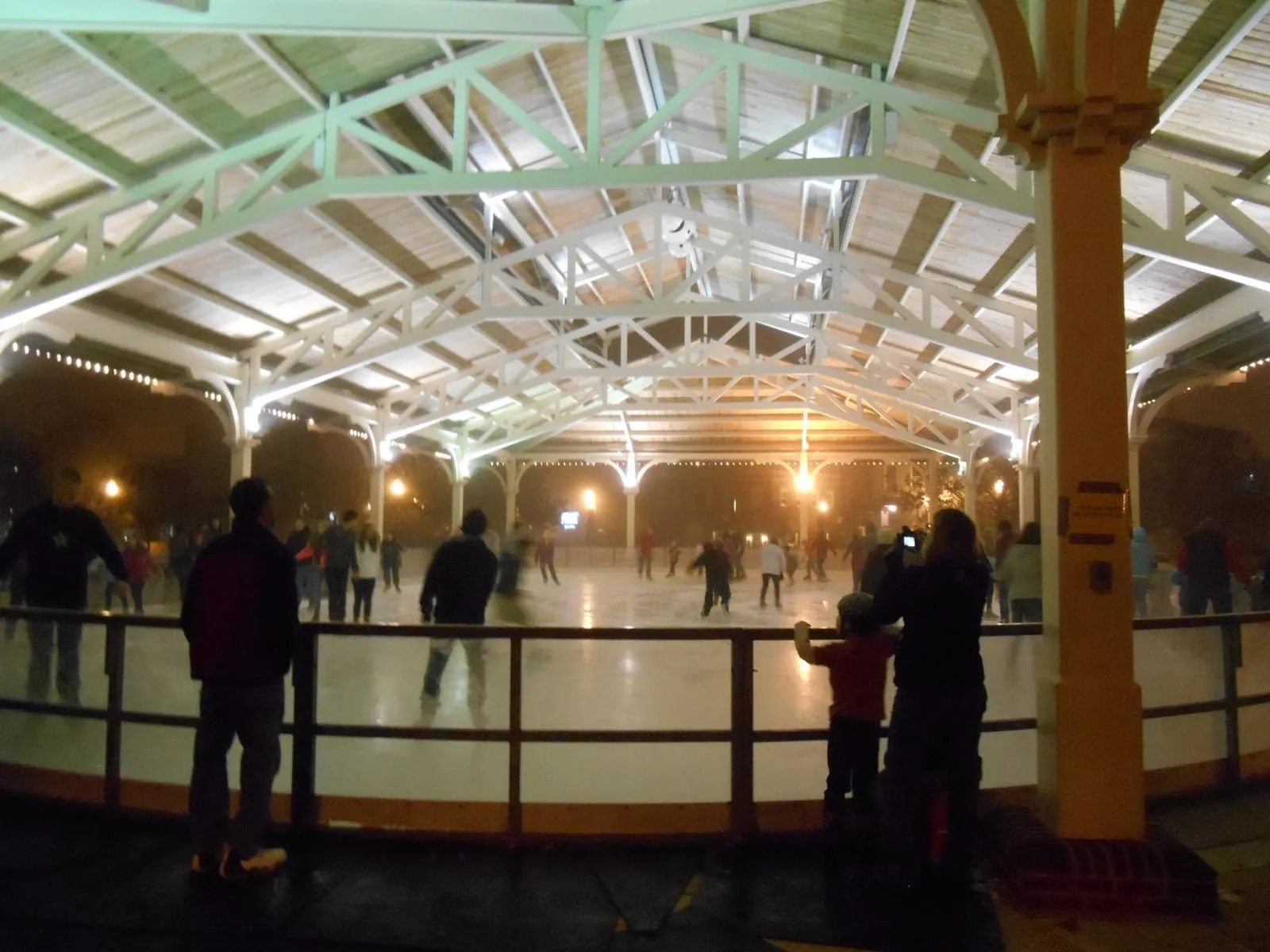 The refrigerated sheet under the open timber-truss roof of the Harris Pavilion at night, with skaters on the ice and onlookers at the boards. — Harris Pavilion Ice Skating