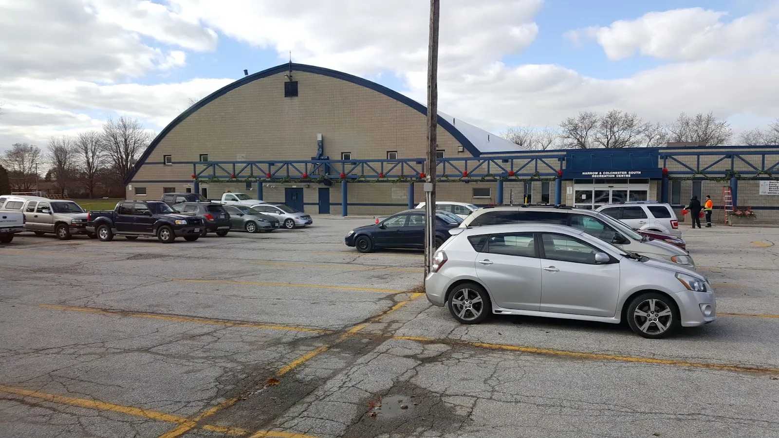 El edificio del estadio con techo curvo detrás de su estacionamiento, con una marquesina de entrada con ribete azul. — Harrow and Colchester South Arena