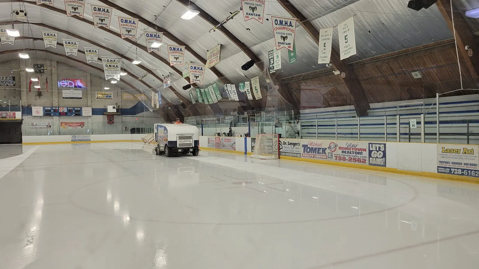 El hielo recién alisado con la máquina al fondo, las vallas con paneles publicitarios y los banderines colgados del techo arqueado. — Harrow and Colchester South Arena